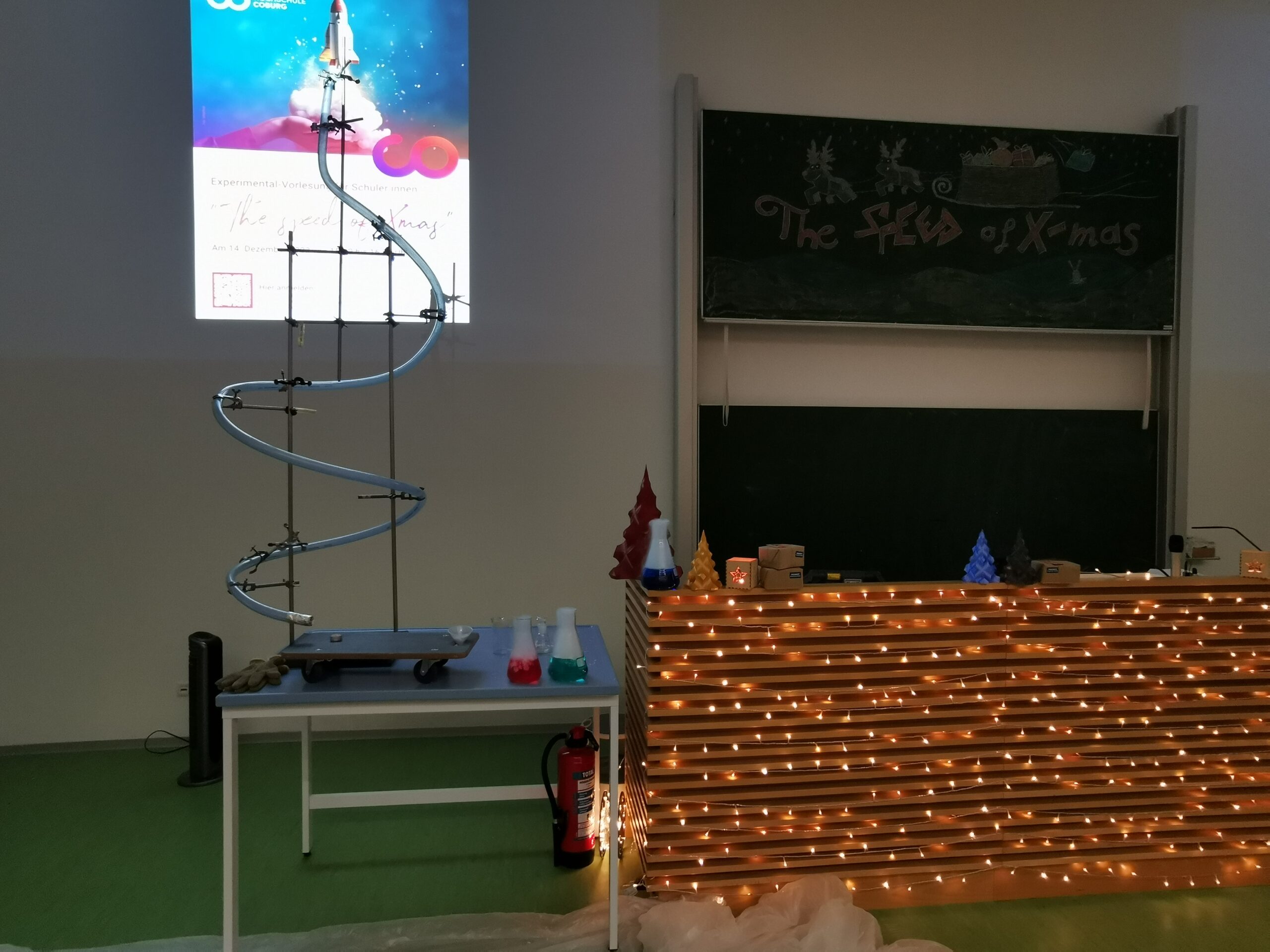 Der Unterrichtsraum der Hochschule Coburg ist festlich für Weihnachten dekoriert und mit einer Präsentationsanlage ausgestattet. Auf dem Tisch steht eine Spiralbahn mit bunten Flaschen, während die Tafel mit Lichtern und festlichen Zeichnungen leuchtet. Eine digitale Projektion beleuchtet die Wand und sorgt für Weihnachtsstimmung.