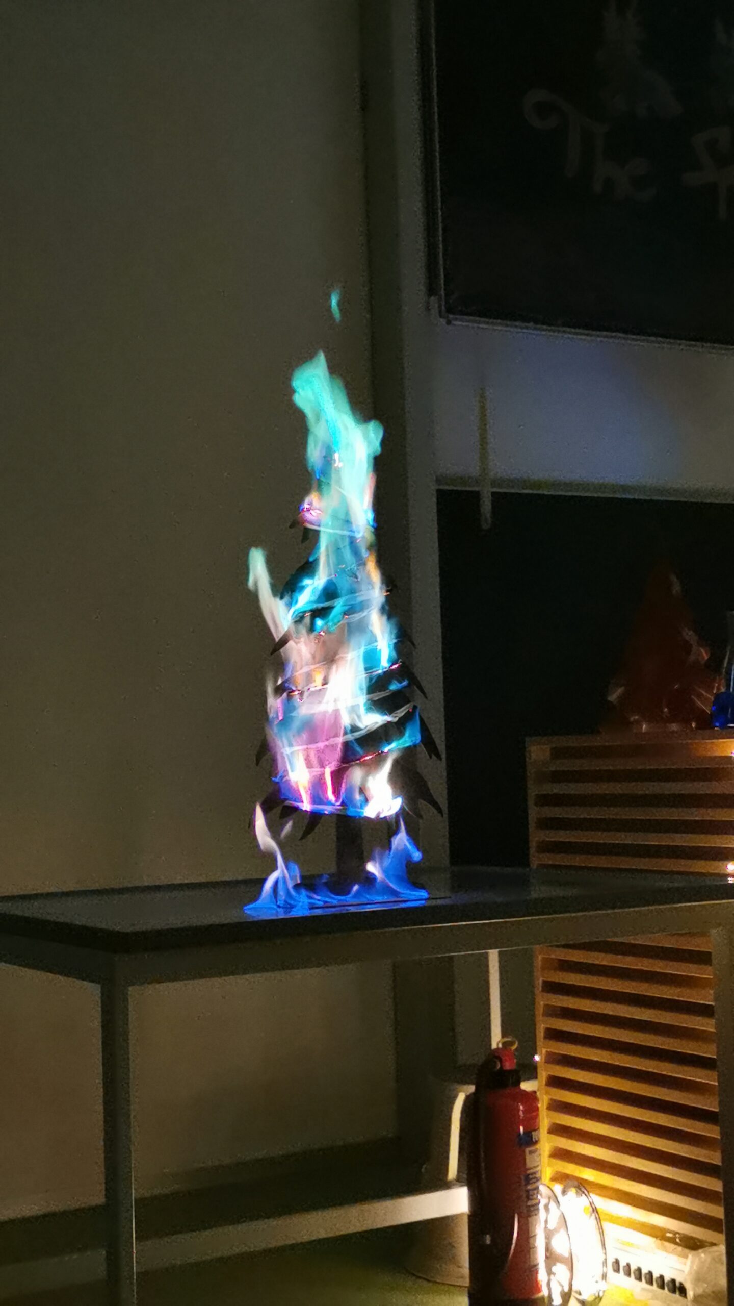 Ein dreieckiger Stapel Materialien wird angezündet, wobei auf einem Metalltisch in der Hochschule Coburg leuchtend blaue, grüne und orange Flammen entstehen. Zur Sicherheit steht ein Feuerlöscher an der Seite.