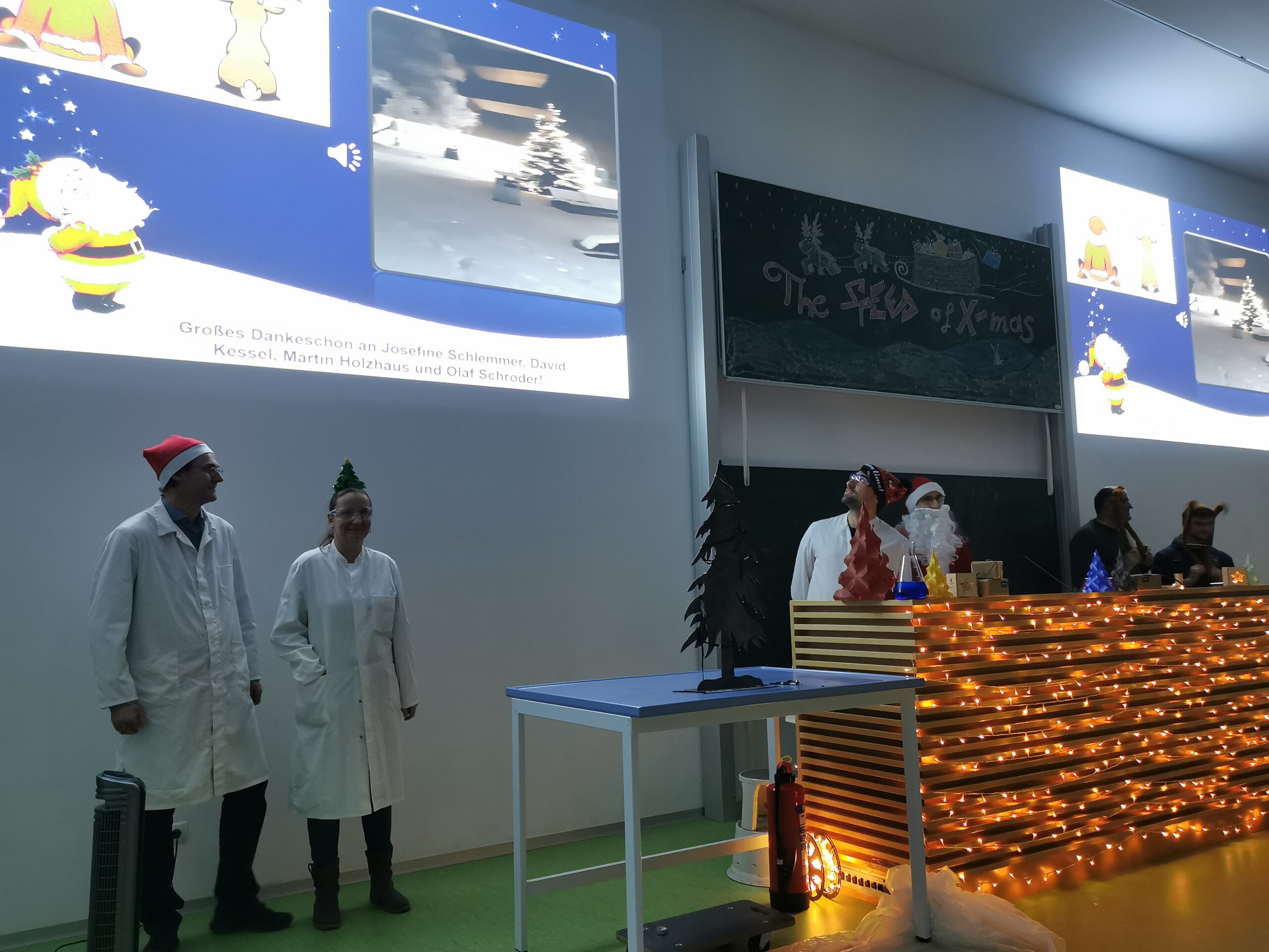 Menschen in Laborkitteln und Weihnachtsmannmützen stehen auf einer weihnachtlich geschmückten Bühne und repräsentieren die Hochschule Coburg. Auf einer Tafel steht „Der Fisch der Weihnacht“, während im Hintergrund eine Präsentation mit Bildern des Weihnachtsmanns und eines Baumes projiziert wird.