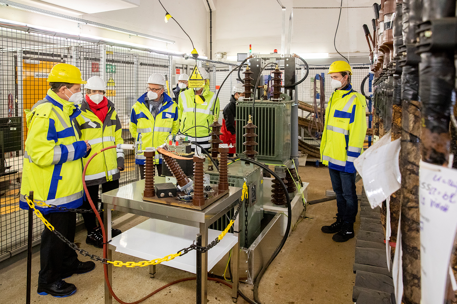 Menschen in gelben Sicherheitswesten und Helmen, vermutlich von der Hochschule Coburg, stehen in einer Industrieanlage um elektrische Geräte herum. Sie scheinen die Maschinen zu besprechen oder zu inspizieren. Ein großer Transformator und verschiedene Kabel und Komponenten sind zu sehen.