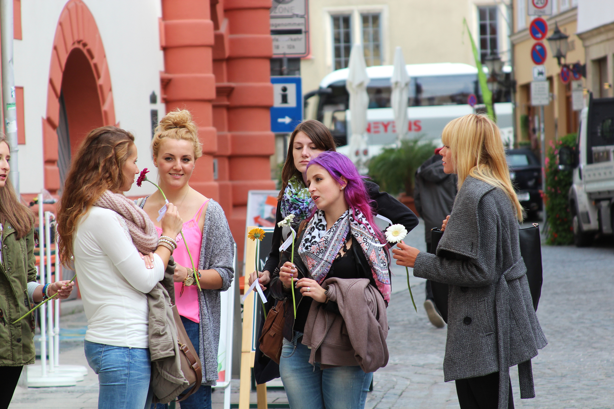 Eine Gruppe von Frauen steht auf einer Kopfsteinpflasterstraße in der Nähe der Hochschule Coburg und unterhält sich. Einige halten Blumen in den Händen. Im Hintergrund sind ein roter Torbogen und ein geparkter Lieferwagen zu sehen. Stadtgebäude und Straßenschilder runden die Szene ab.