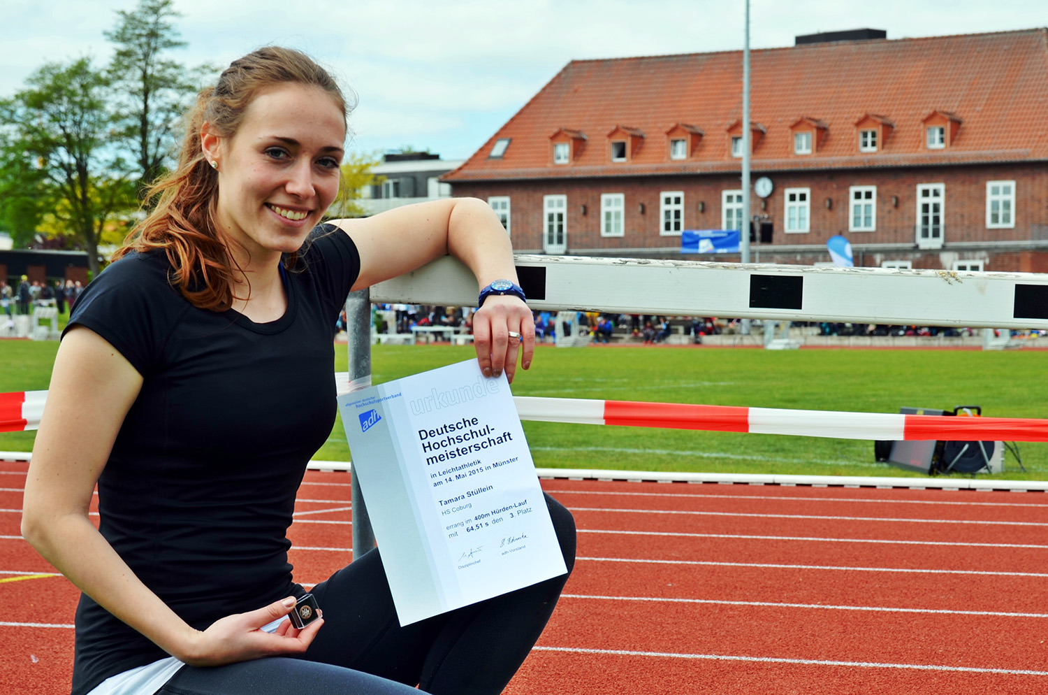 Eine Frau in Sportkleidung kniet lächelnd auf einer Leichtathletikstrecke und hält ein Zertifikat der Hochschule Coburg in der Hand. Im Hintergrund sieht man ein Backsteingebäude mit rotem Dach und in der Ferne eine Gruppe von Menschen.