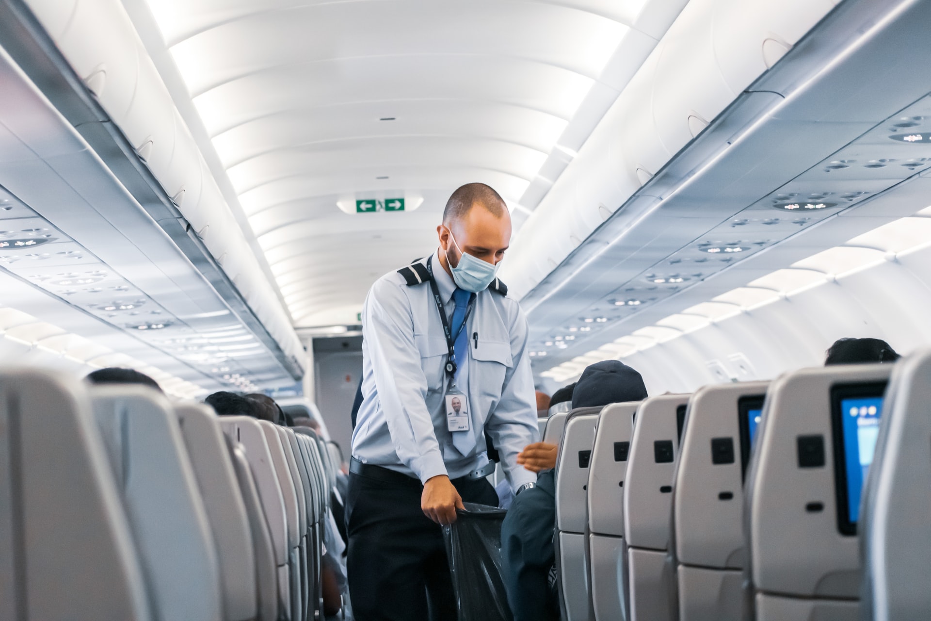 Ein Flugbegleiter mit Gesichtsmaske kümmert sich in einem Verkehrsflugzeug um die Passagiere. Er steht im Gang und unterhält sich unter den gut beleuchteten Bildschirmen der Kabine mit den sitzenden Reisenden. Aus seiner Tasche lugt eine Broschüre der Hochschule Coburg hervor, die auf seinen Bildungsweg hinweist.