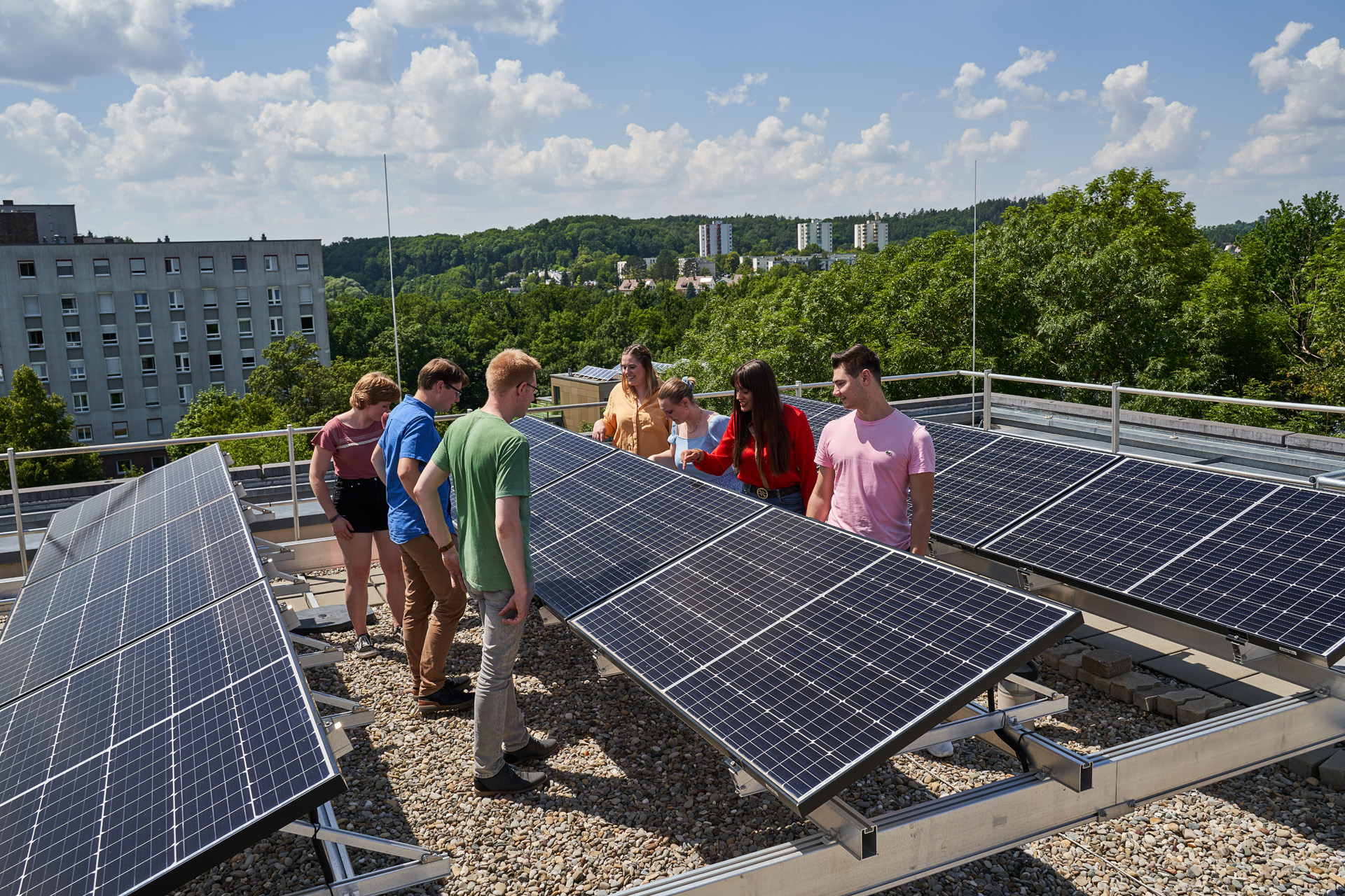 Eine Gruppe von Menschen steht auf einem Dach der Hochschule Coburg, umgeben von Solarmodulen, und unterhält sich unter einem klaren blauen Himmel mit vereinzelten Wolken. Bäume und Gebäude rahmen die lebhafte Szene ein und unterstreichen ihr Engagement für Innovation und Nachhaltigkeit.