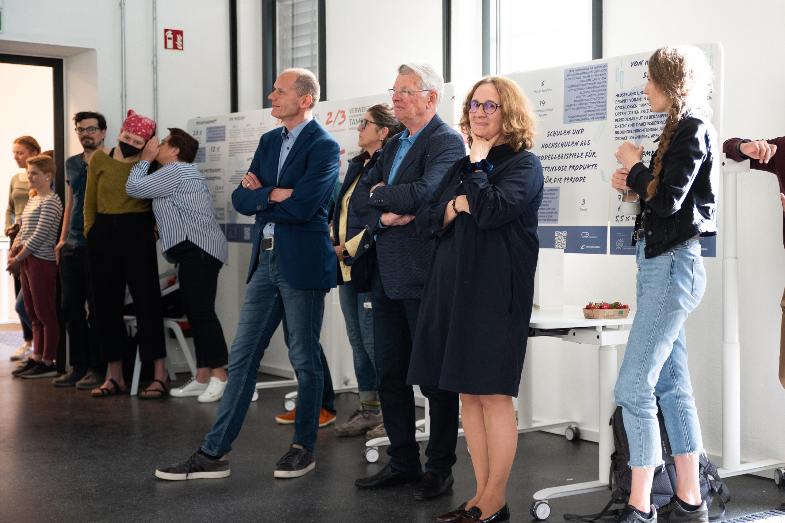 Eine Gruppe von Menschen steht in einem hell erleuchteten Raum der Hochschule Coburg und betrachtet Präsentationen auf Postern. Einige lehnen an einer Wand, andere stehen mit verschränkten Armen da. Die Atmosphäre wirkt konzentriert und aufmerksam.