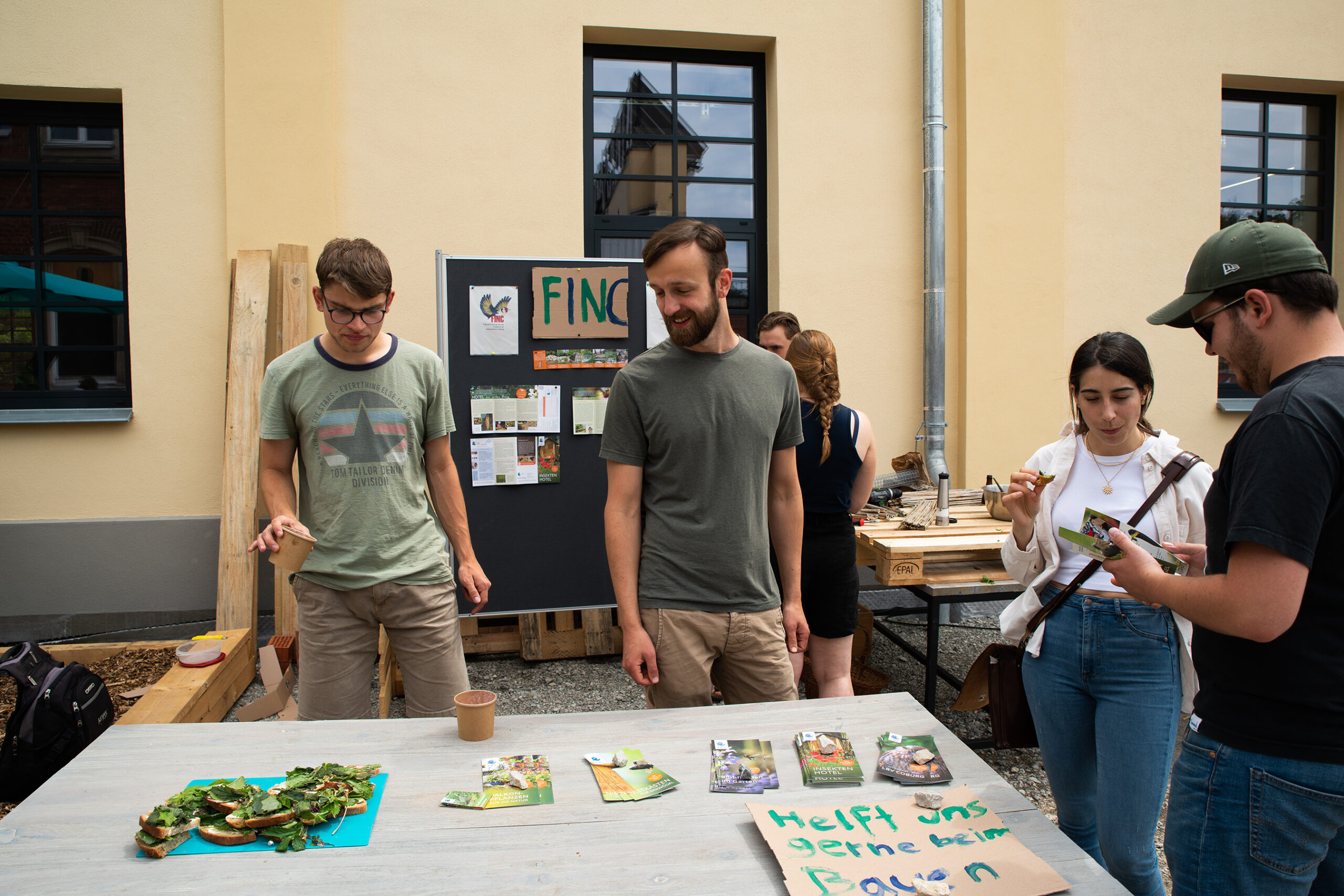 Vor einem Gebäude der Hochschule Coburg steht eine Gruppe von Menschen um einen Tisch mit Pflanzen und Broschüren. Eine Person zeigt auf die Gegenstände auf dem Tisch, eine andere macht ein Foto davon. Daneben steht eine Pinnwand mit Informationen unter der Überschrift „FINCA“.