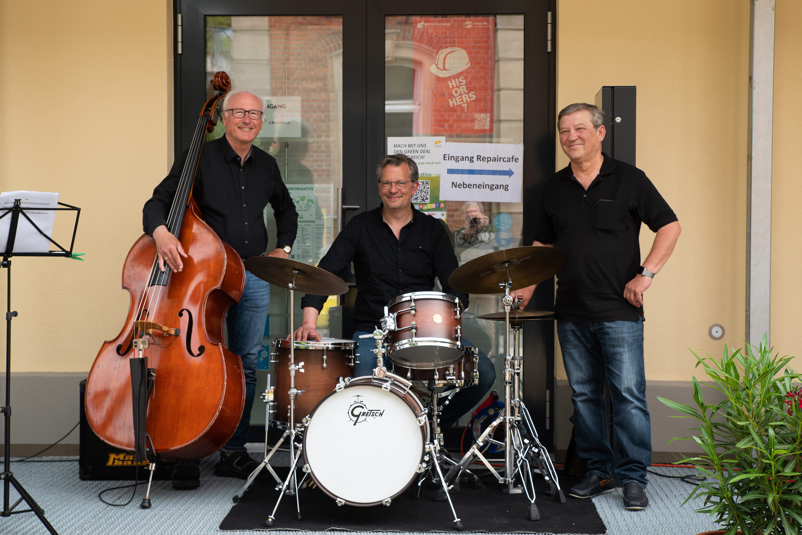 Drei Männer in schwarzen Hemden posieren mit Musikinstrumenten vor einem Gebäude der Hochschule Coburg. Einer steht mit einem Kontrabass, ein anderer sitzt hinter einem Schlagzeug und der dritte steht daneben. Im Vordergrund sind ein Notenständer und eine Pflanze zu sehen.