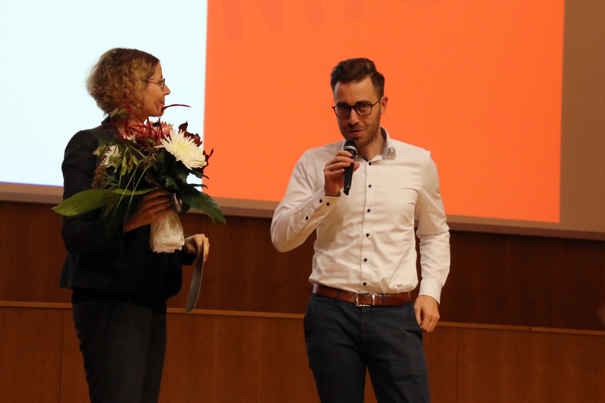 Ein Mann im weißen Hemd hält auf der Bühne ein Mikrofon, neben ihm steht eine Frau in schwarzer Jacke mit einem Blumenstrauß. Sie befinden sich in einem geräumigen Raum mit der für die Hochschule Coburg typischen rot-weißen Kulisse.