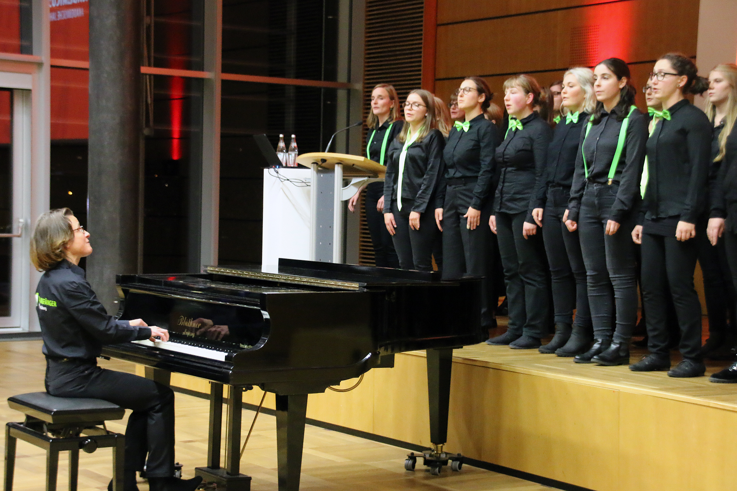 Eine Gruppe von Chorsängern der Hochschule Coburg, in schwarzen Kostümen mit grünen Fliegen, tritt auf einer Bühne auf. Ein Pianist, ebenfalls in Schwarz, begleitet sie auf einem Flügel. Die Kulisse scheint ein moderner, gut beleuchteter Hörsaal zu sein.