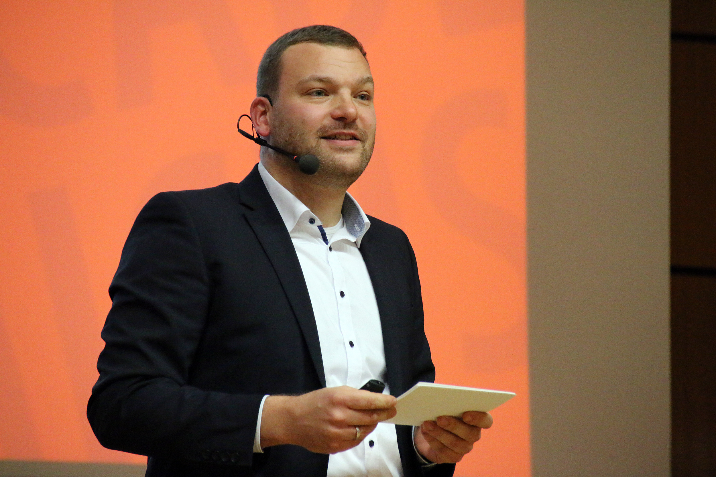 Ein Mann im Anzug, der ein Headset-Mikrofon trägt, hält einen Notizblock, während er vor einem orangefarbenen Hintergrund an der Hochschule Coburg eine spannende Rede hält.