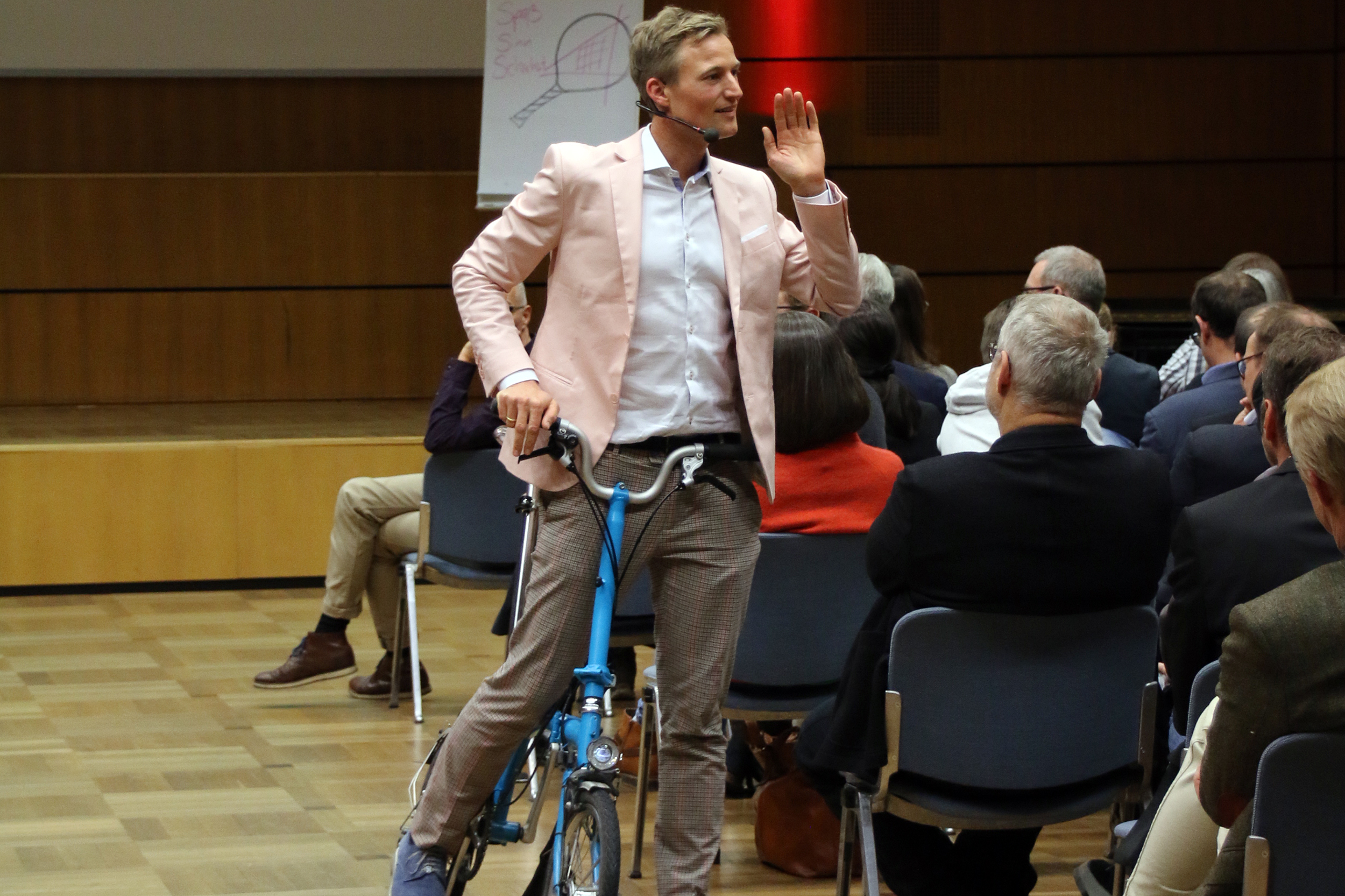 Ein Mann in einem rosa Blazer steht mit einem Fuß auf einem kleinen blauen Faltrad und spricht in ein Mikrofon, während er sich an ein Publikum wendet, das in Reihen im Hörsaal der Hochschule Coburg sitzt.