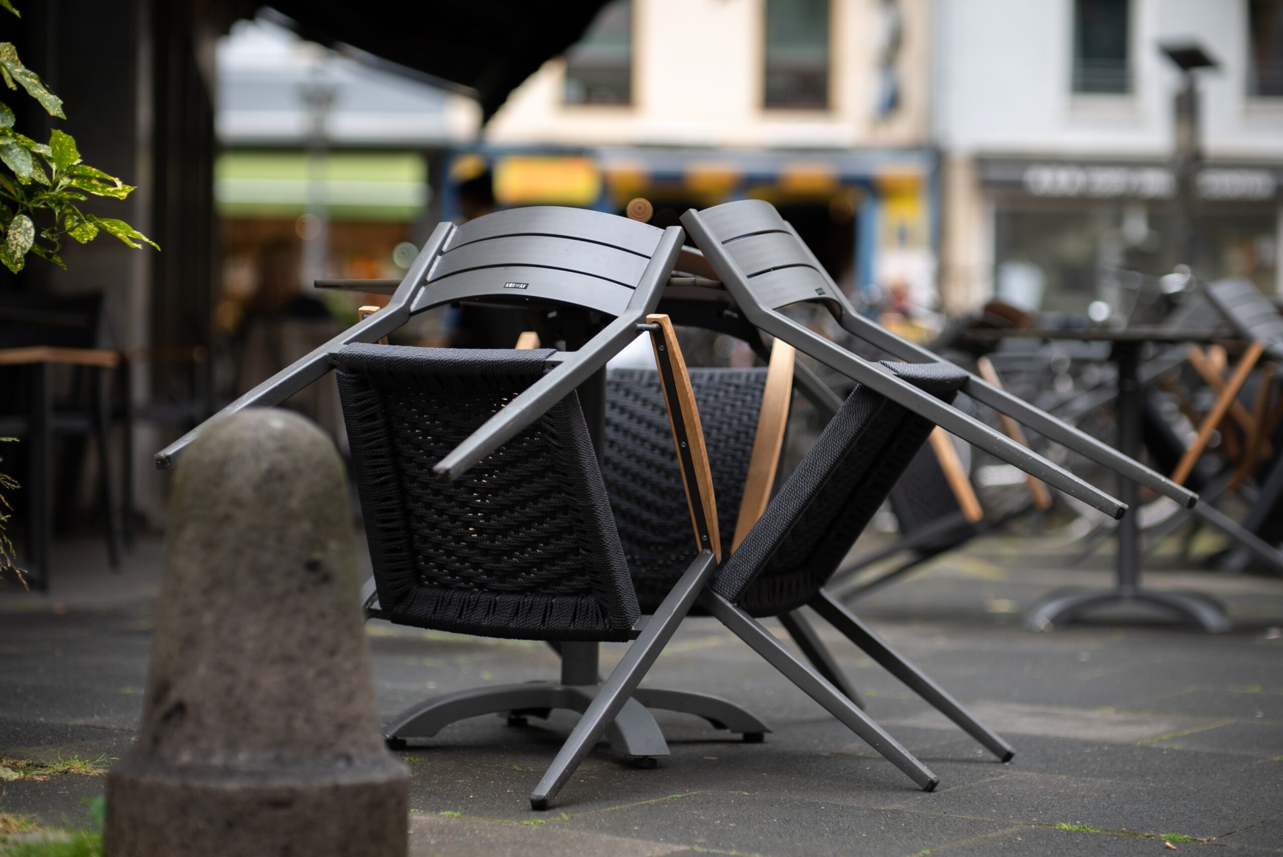 Gestapelte schwarze Terrassenstühle auf der Terrasse eines Straßencafés in der Nähe der Hochschule Coburg, im Hintergrund sind verschiedene Gebäude verschwommen. Im Vordergrund steht ein Steinpoller, der zu einer ruhigen und bewölkten Atmosphäre beiträgt.