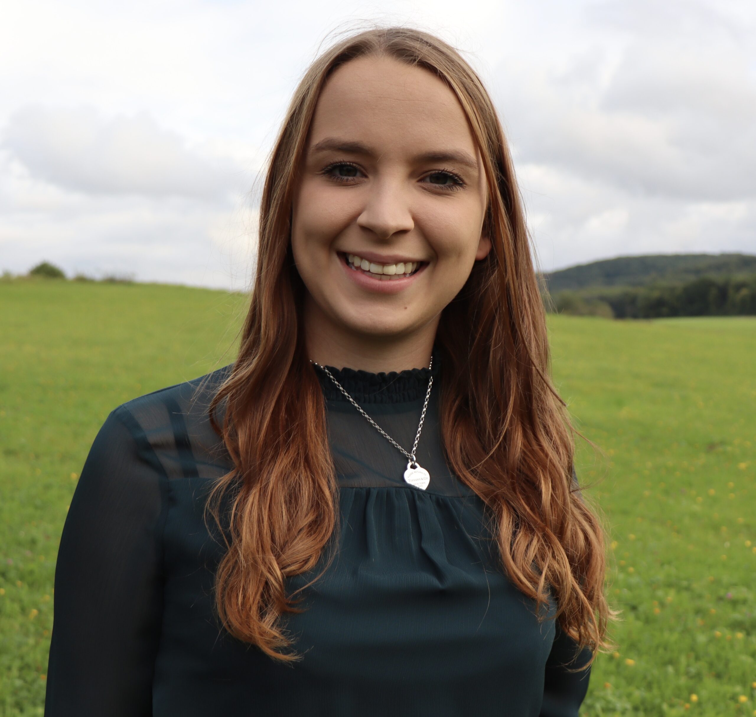 Eine Person mit langen braunen Haaren und einer Halskette steht lächelnd auf einer grünen Wiese unter dem bewölkten Himmel in der Nähe der Hochschule Coburg.