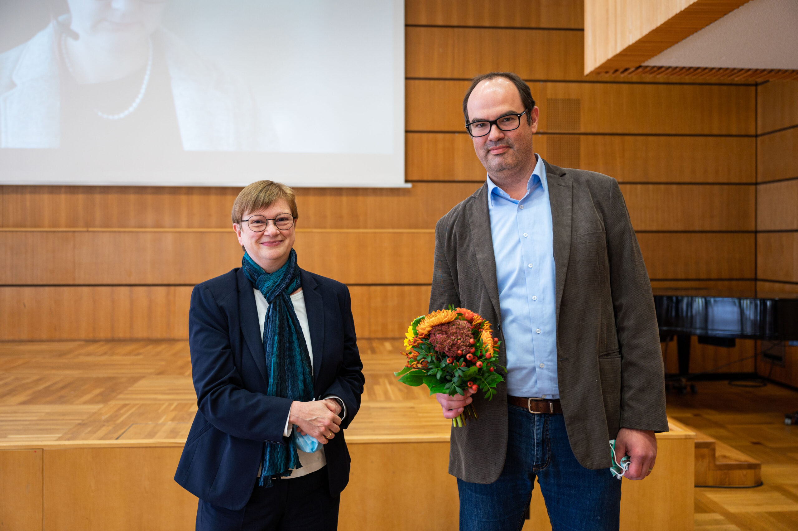 Zwei Menschen stehen in einem Hörsaal der Hochschule Coburg. Die rechte Person trägt Brille und Jackett und hält einen Blumenstrauß in der Hand. Die linke Person ist mit Anzug und Schal bekleidet. Holzvertäfelungen und ein Paravent unterstreichen das edle Ambiente.