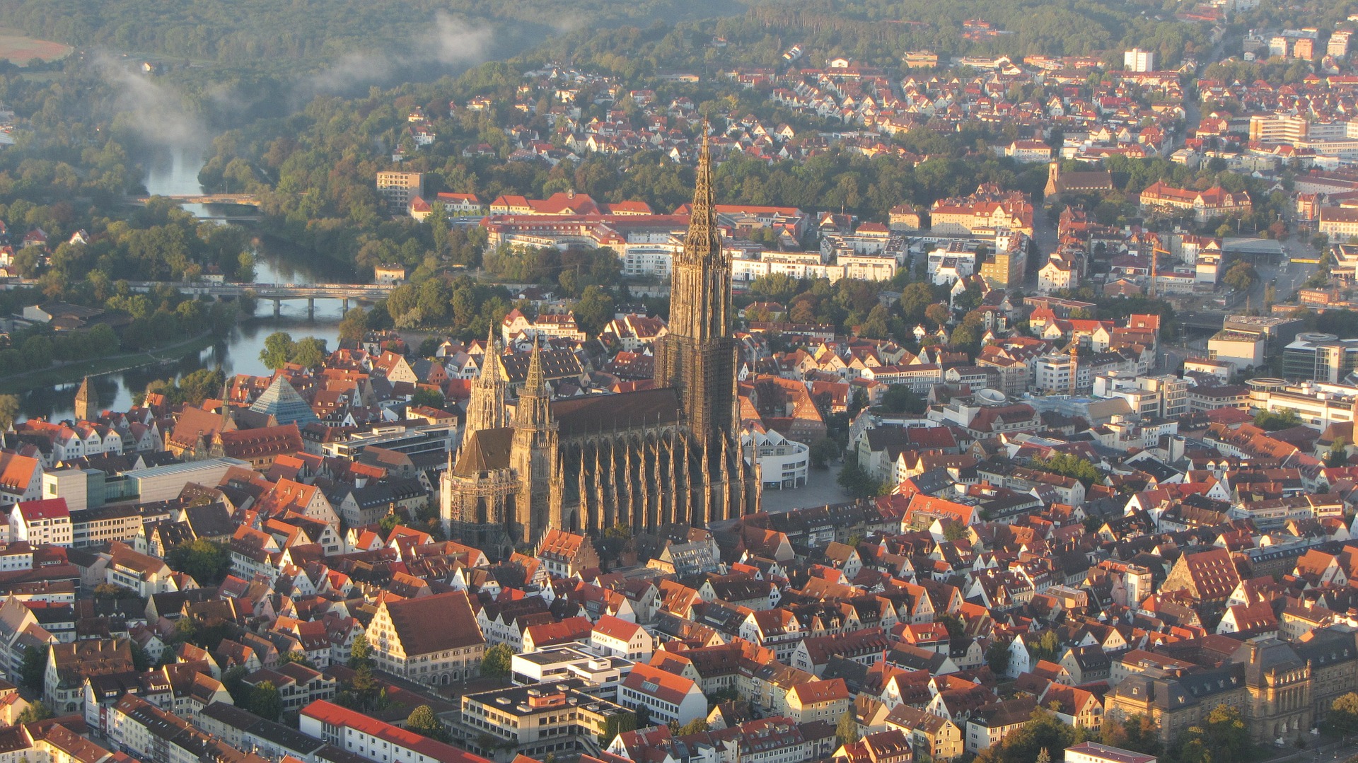 Eine Luftaufnahme einer Stadt zeigt eine markante gotische Kathedrale mit einem hohen Turm in der Mitte. Die Umgebung, in deren Nähe sich die Hochschule Coburg befindet, zeichnet sich durch zahlreiche Gebäude mit roten Dächern, Straßen und Grünflächen aus, alles vor der Kulisse eines gewundenen Flusses.