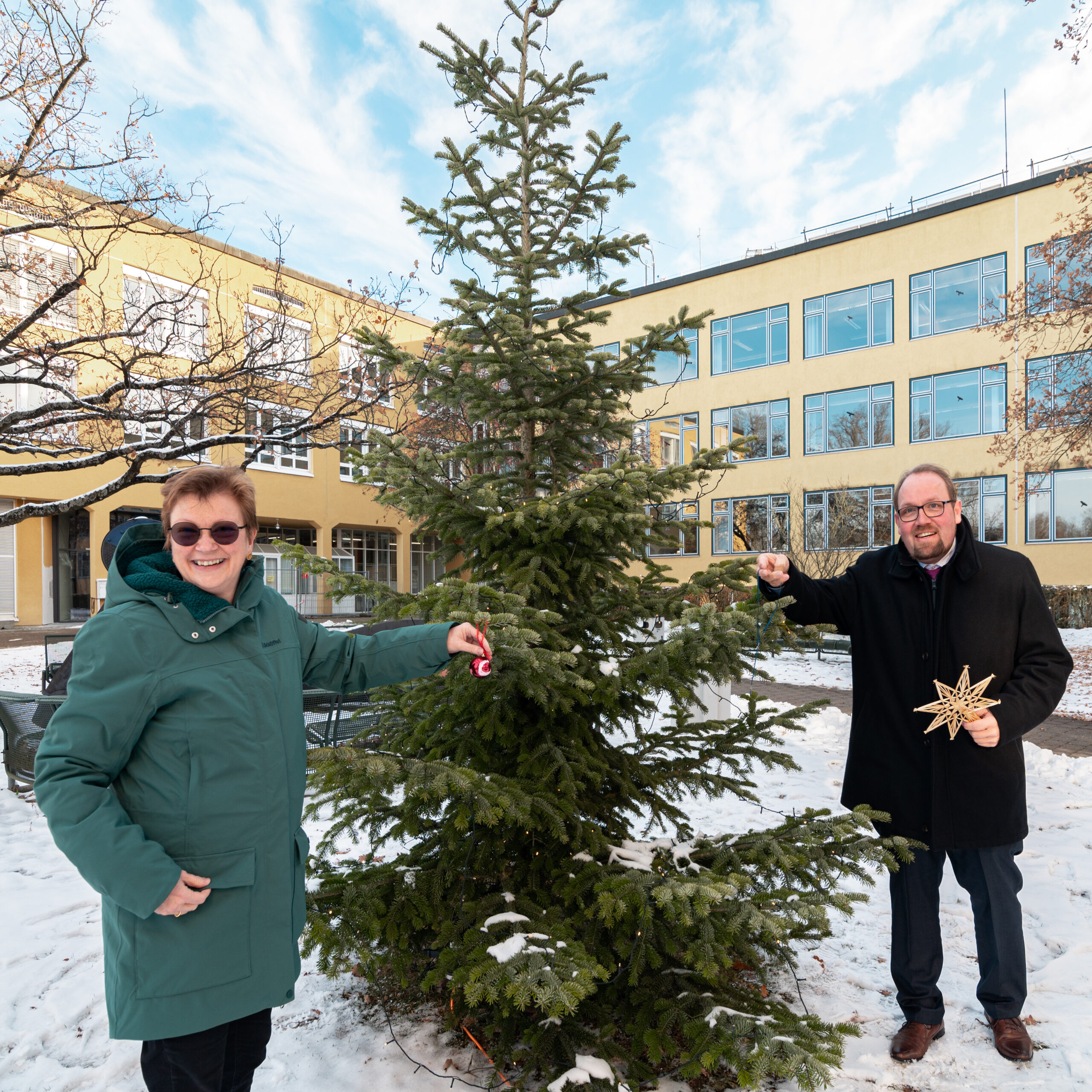 Zwei Personen schmücken einen kleinen Weihnachtsbaum im Freien vor der Hochschule Coburg mit Ornamenten. Die Person links trägt einen grünen Mantel und die Person rechts, in einem schwarzen Mantel, hält einen Stern. Im Hintergrund steht stolz ein gelbes Gebäude, während der Boden mit Schnee bedeckt ist.