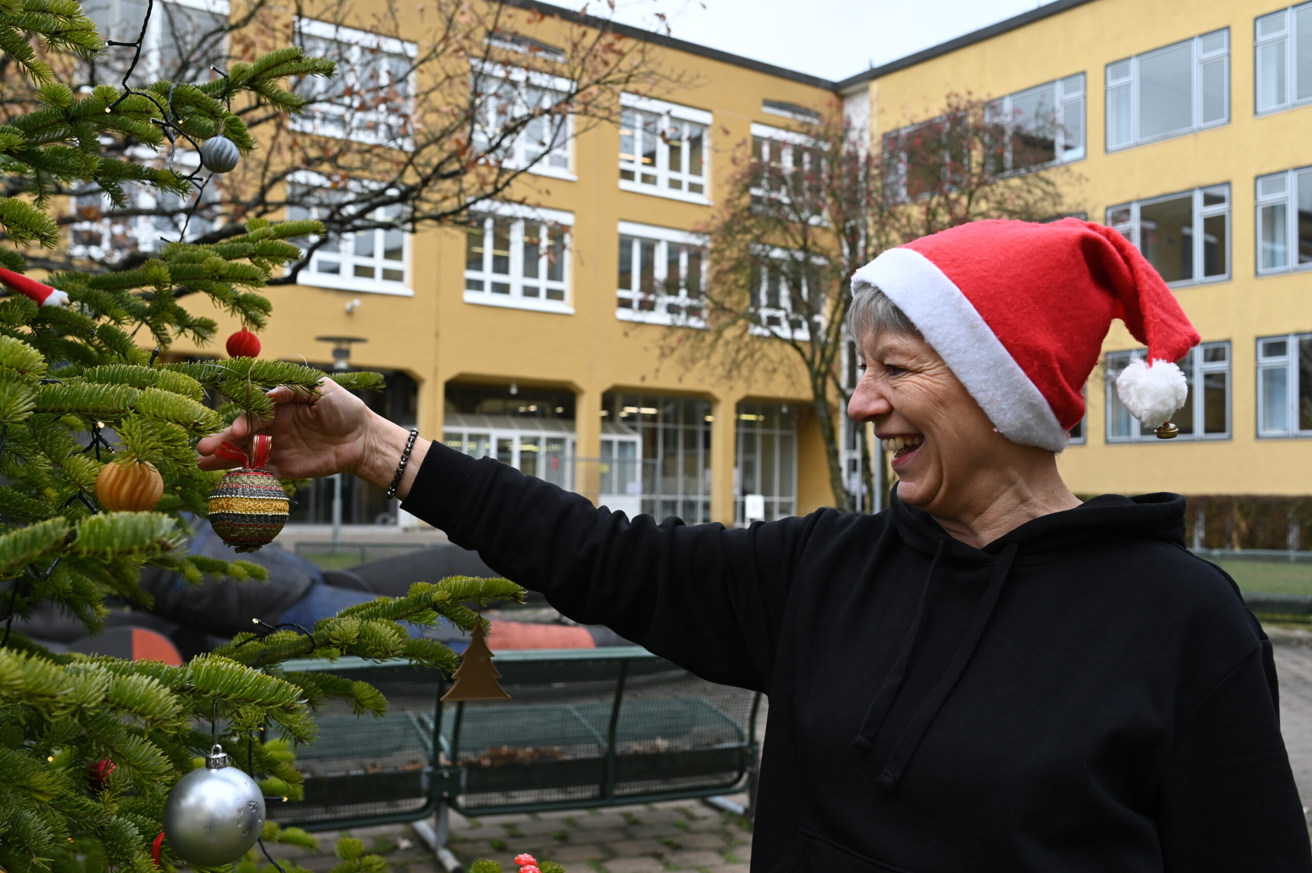 Eine ältere Frau mit Weihnachtsmannmütze schmückt einen Weihnachtsbaum vor der Hochschule Coburg und hängt voller Freude Weihnachtsschmuck auf. Sie lächelt herzlich und trägt einen schwarzen Kapuzenpullover. Im Hintergrund steht ein gelbes Gebäude mit bezaubernden weiß gerahmten Fenstern.