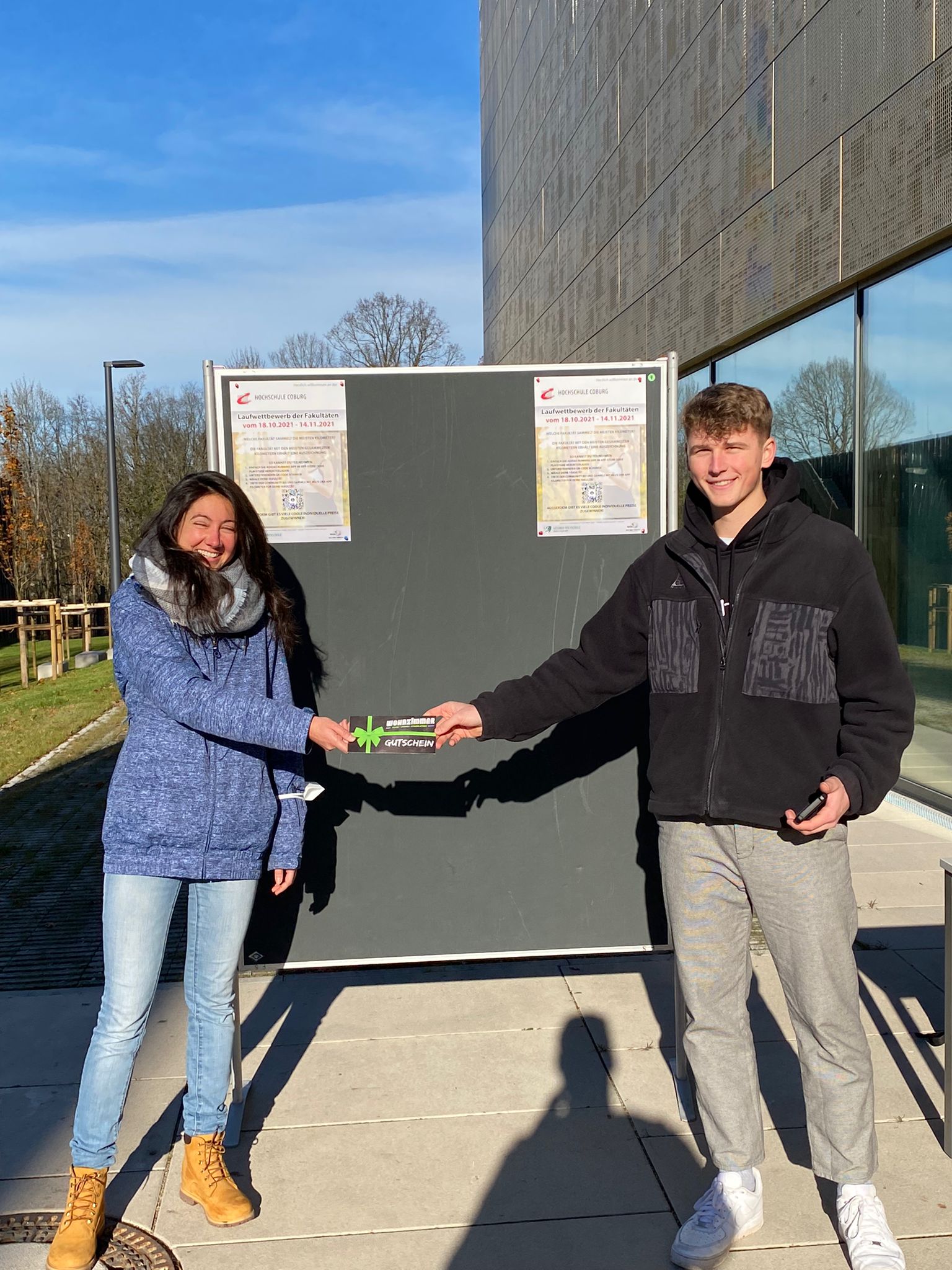 Zwei Personen stehen an einem sonnigen Tag im Freien vor einem schwarzen Brett an der Hochschule Coburg. Sie lächeln und schütteln sich die Hände, und einer hält einen kleinen rechteckigen Gegenstand in der Hand. Beide tragen lässige Winterkleidung, darunter Jacken und Jeans.