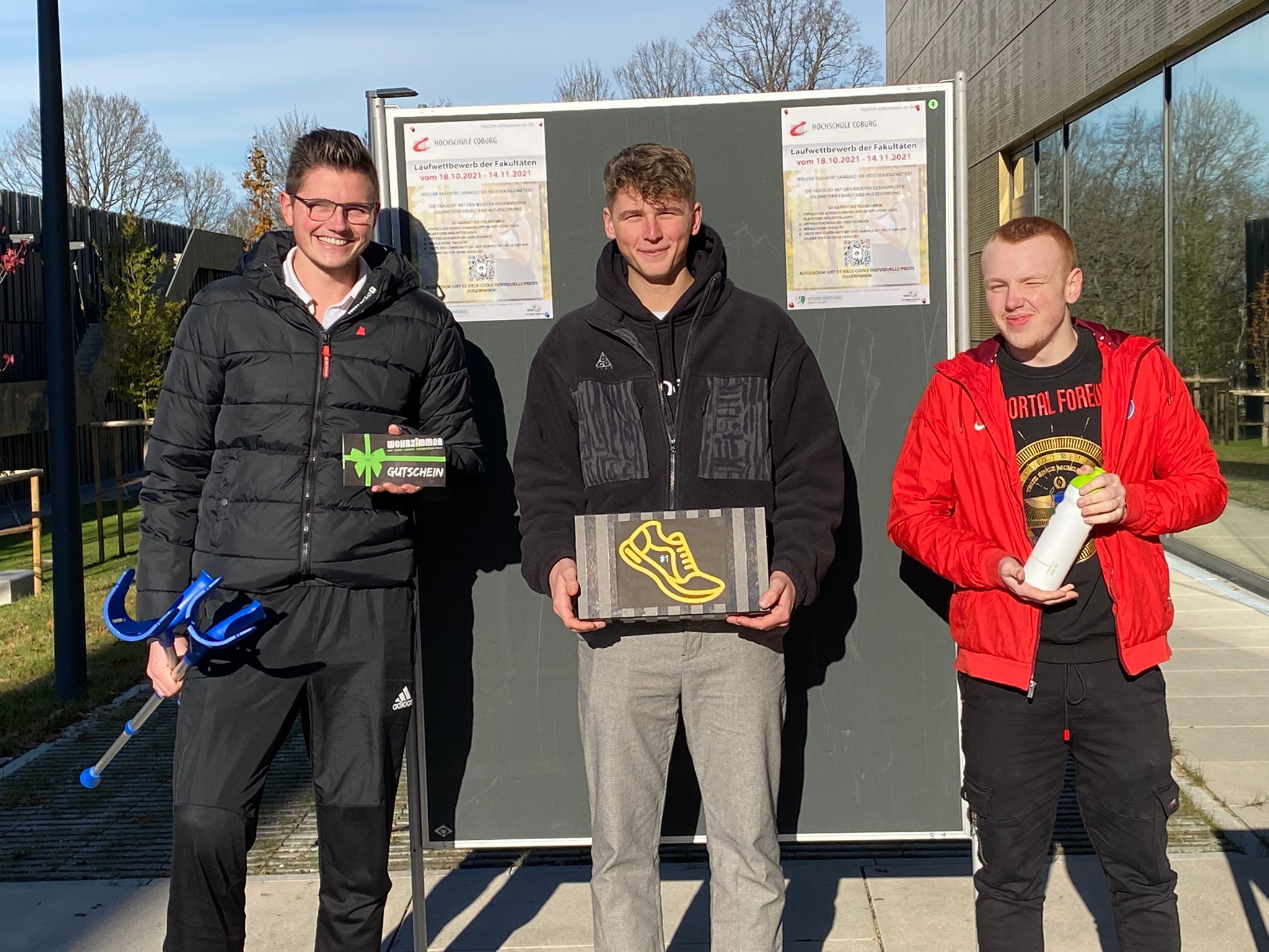 Drei junge Männer stehen an einem sonnigen Tag vor der Hochschule Coburg, lächeln und halten verschiedene Gegenstände in den Händen. Der erste hält ein Fahrradwerkzeug, der zweite eine Schachtel mit einem gelben Schuhbild und der dritte eine weiße Flasche. Im Hintergrund sind ein Gebäude und eine Informationstafel zu sehen.