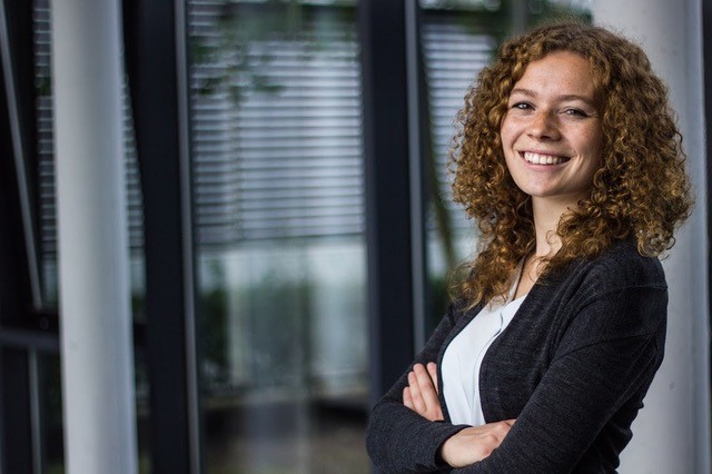 Eine Person mit lockigem Haar steht mit verschränkten Armen in einem Ambiente der Hochschule Coburg und lächelt herzlich. Sie trägt eine schwarze Jacke und ein weißes Hemd. Im Hintergrund sind große Fenster mit Jalousien zu sehen, die eine moderne und professionelle Atmosphäre schaffen.