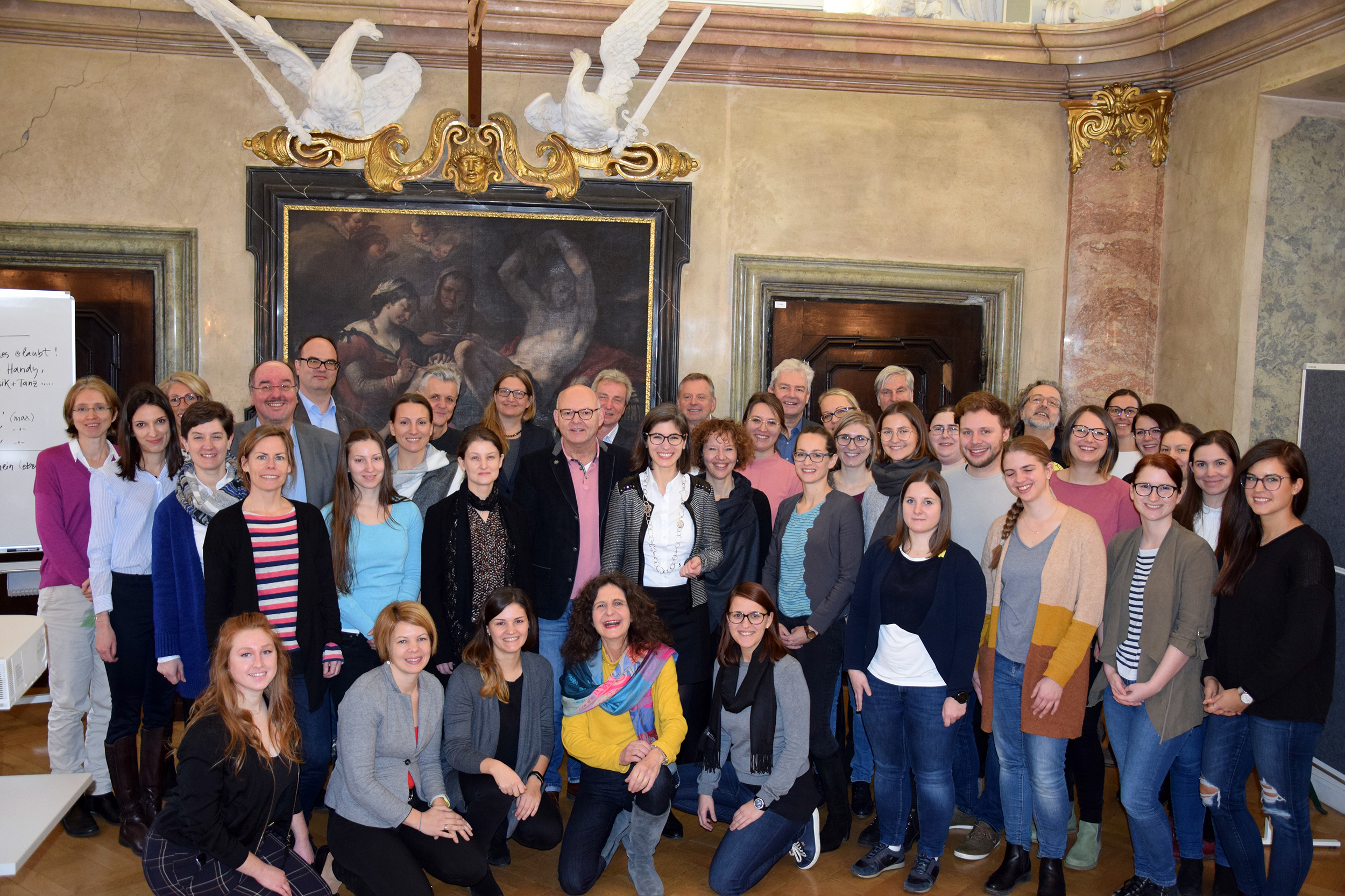 Eine große Gruppe von Menschen, vermutlich von der Hochschule Coburg, posiert für ein Foto in einem dekorierten Raum mit einem Gemälde und Skulpturen im Hintergrund. In Freizeit- und Businesskleidung stehen sie eng beieinander und lächeln strahlend in die Kamera.