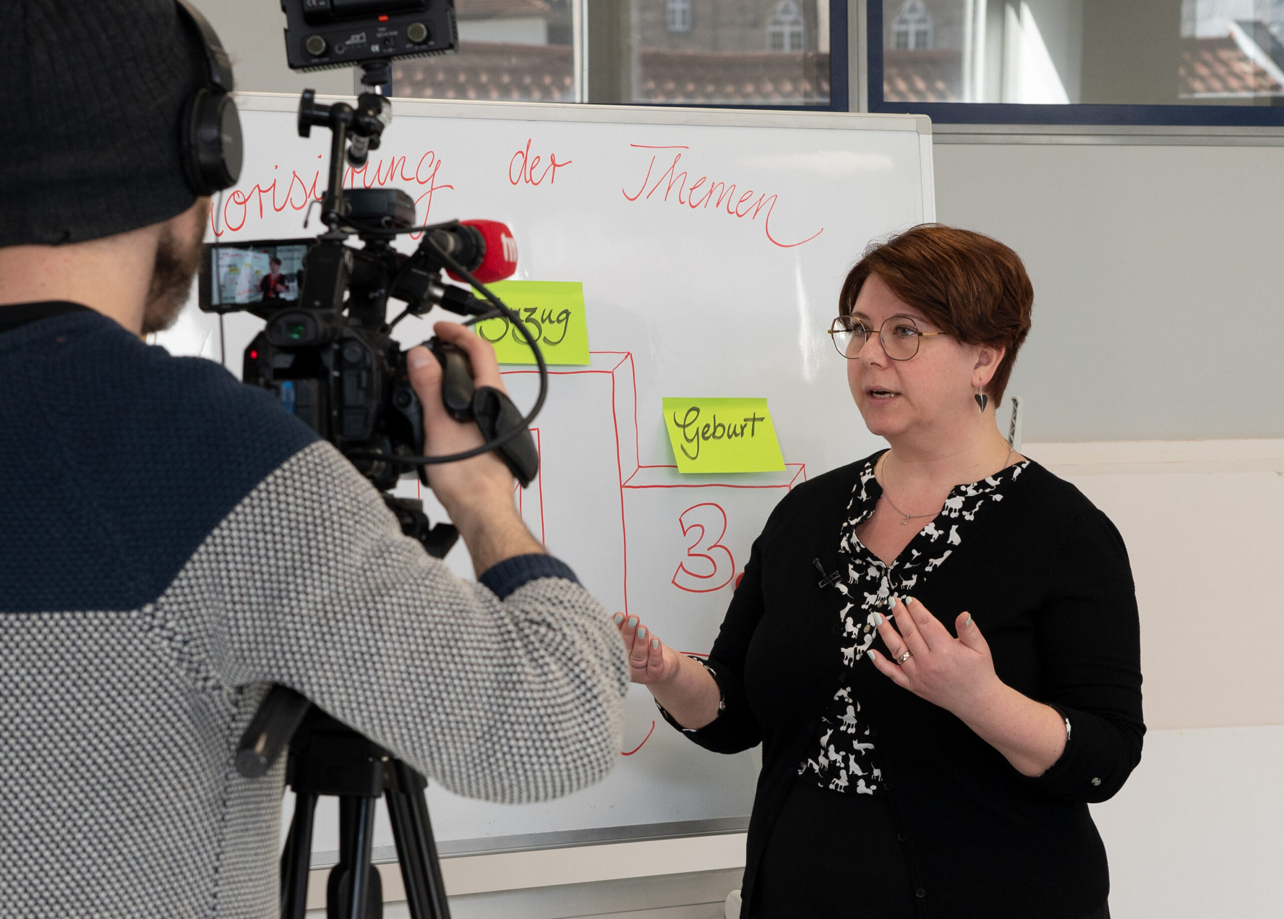 Eine Frau mit Brille steht vor einem Whiteboard an der Hochschule Coburg, spricht angeregt und gestikuliert mit ihren Händen. Währenddessen hält ein Mann mit einer Beaniemütze den Moment auf Video fest. Die akademische Atmosphäre ist lebendig und konzentriert.