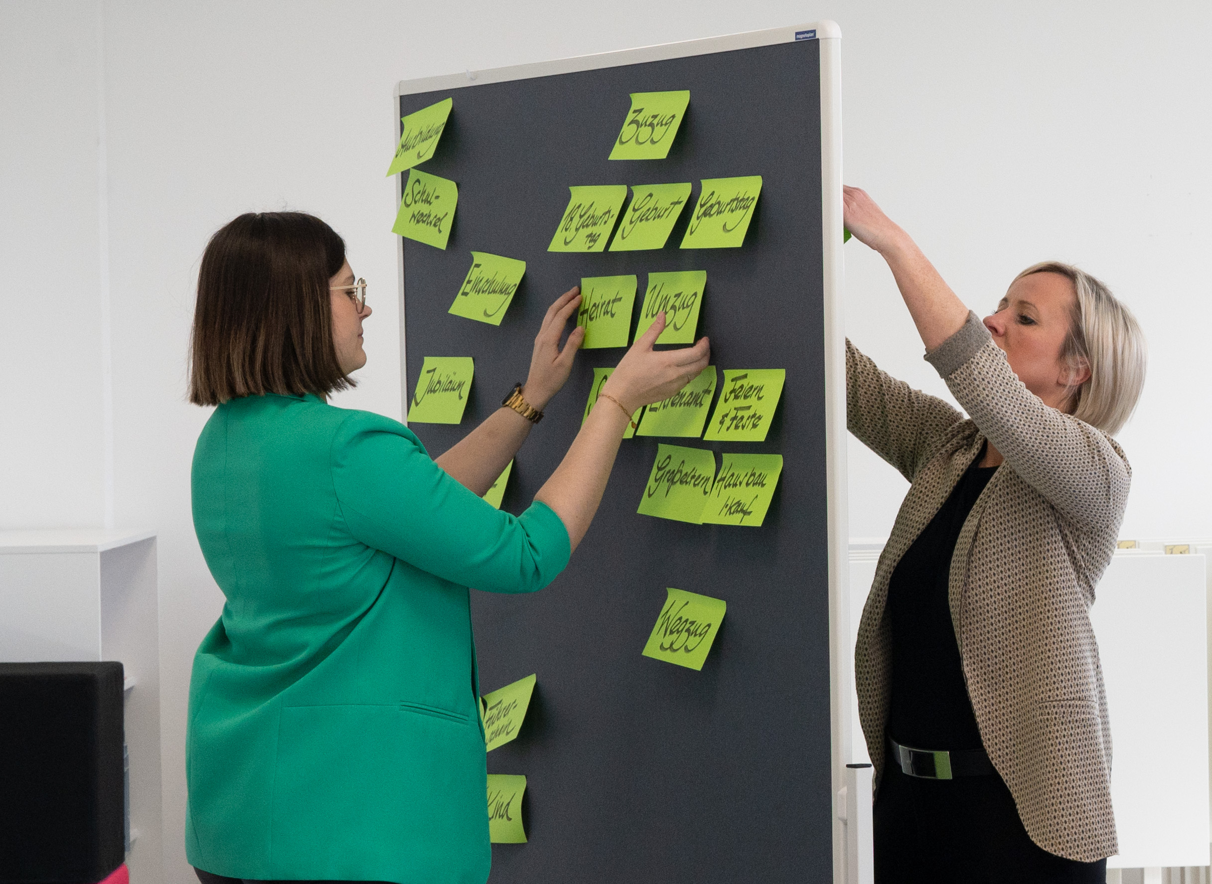 Two people are placing sticky notes with handwritten words on a large board at Hochschule Coburg. The bright green notes contrast the minimalist, well-lit room as both individuals appear focused on organizing their ideas.