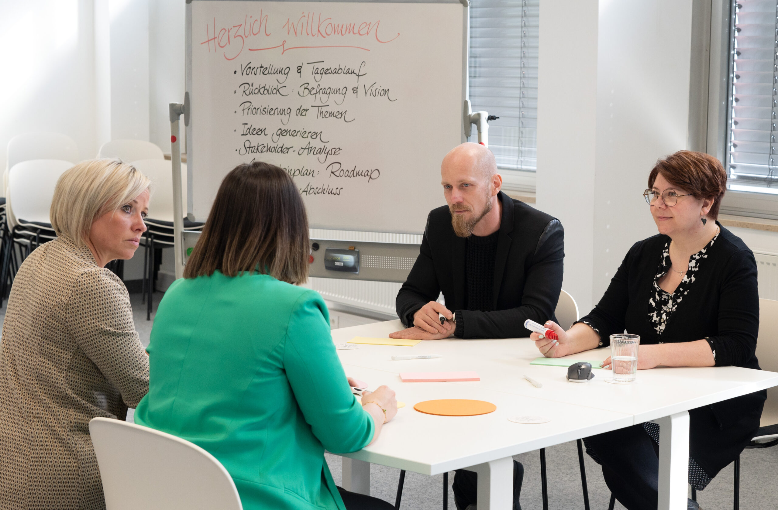 Four people sit around a white table in a meeting room at Hochschule Coburg. A whiteboard with notes in German stands behind them. They appear to be engaged in a discussion, with papers and pens in front of them.