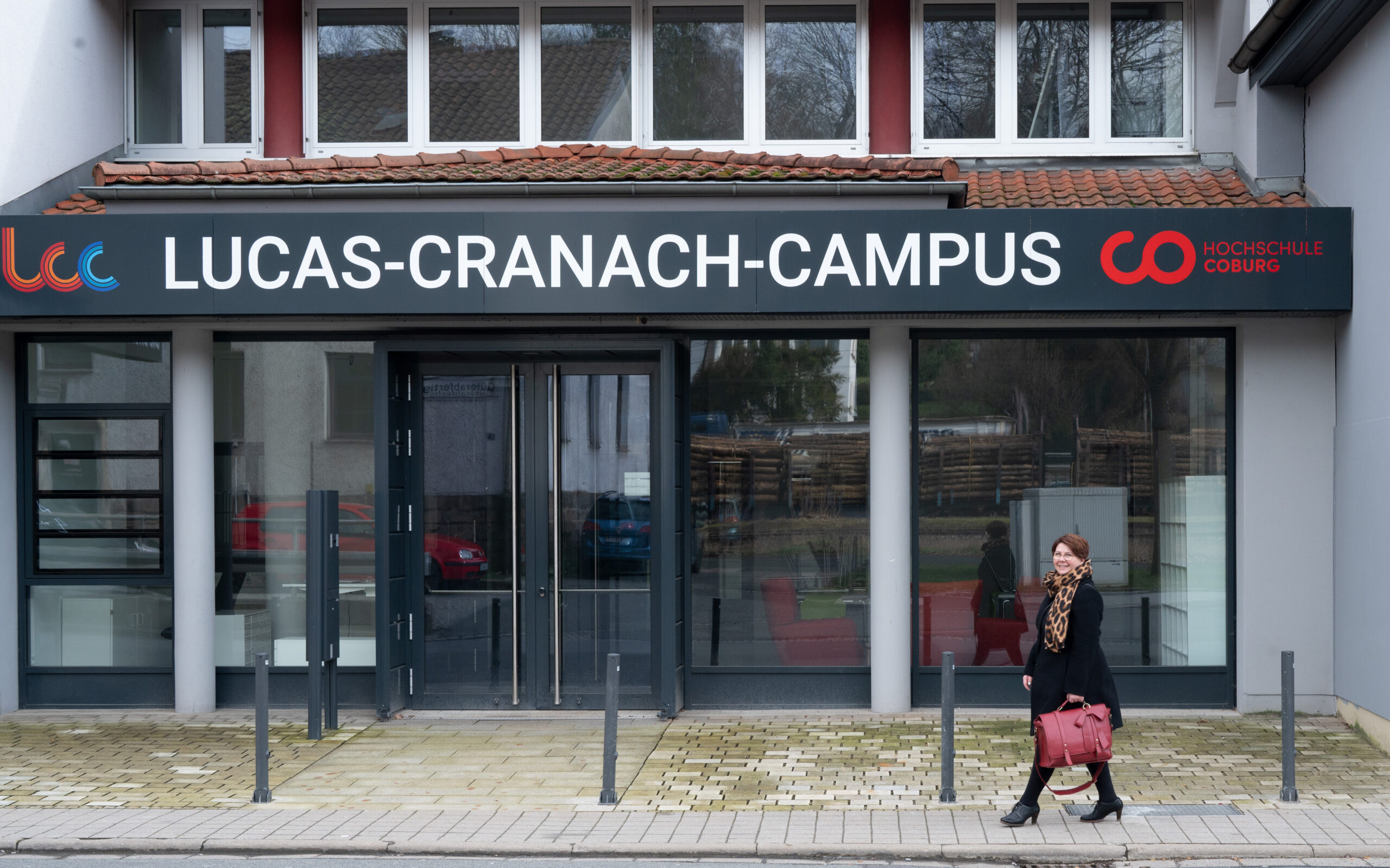 A woman with a red handbag walks past the entrance of Lucas-Cranach-Campus at Hochschule Coburg. The building features large glass doors, a sign showcasing the campus name and logo, and a few bollards strategically placed near the entrance.