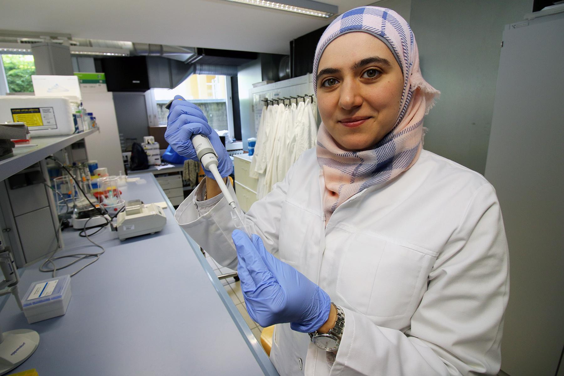 Eine Wissenschaftlerin der Hochschule Coburg, die Hijab, einen weißen Laborkittel und blaue Handschuhe trägt, benutzt im Labor eine Pipette. Im Hintergrund hängen Regale mit Geräten und Laborkitteln, während sie in die Kamera lächelt.