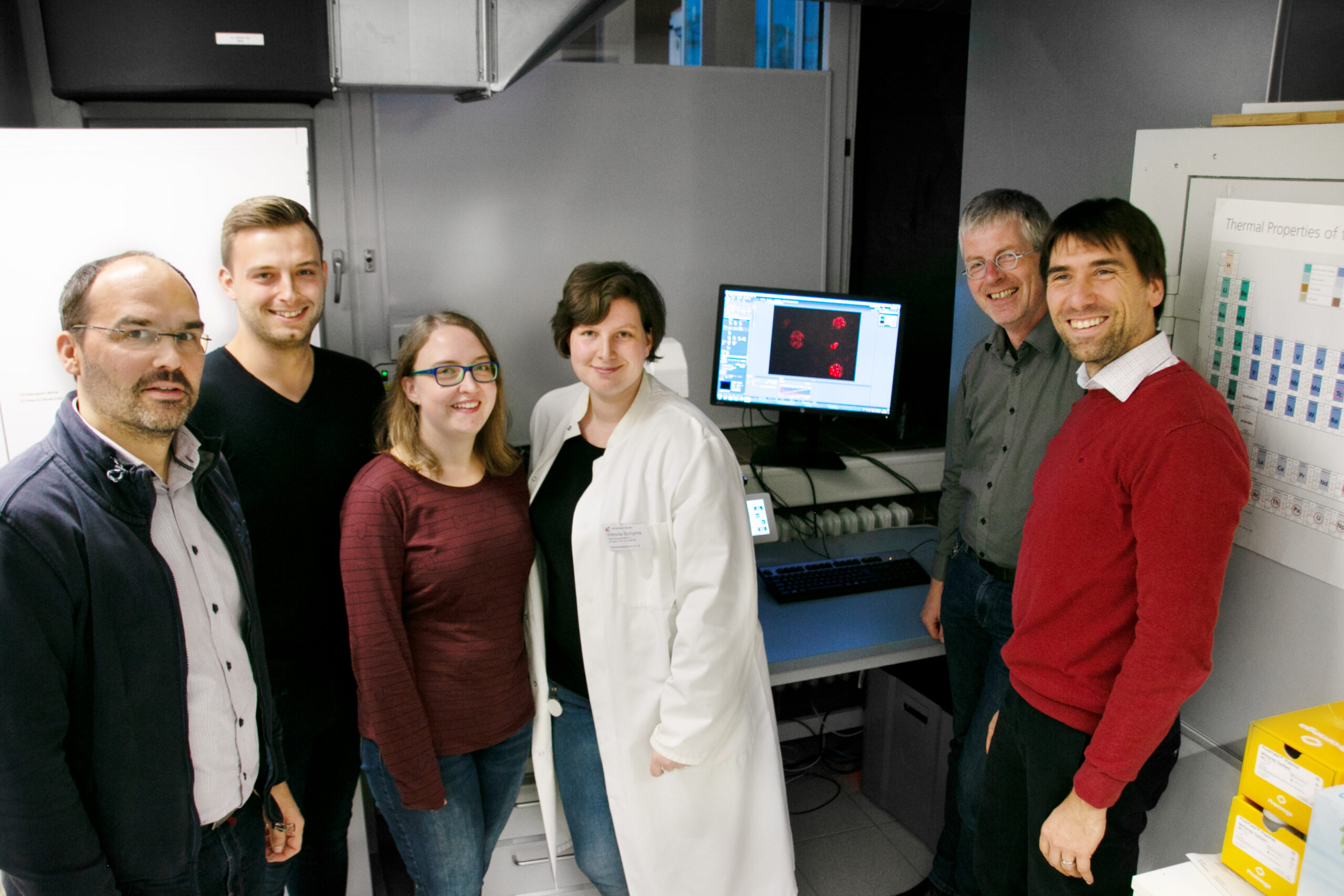 Eine Gruppe von sieben Personen steht in einem Labor der Hochschule Coburg und lächelt in die Kamera. Eine Person trägt einen weißen Laborkittel. Im Hintergrund zeigt ein Computermonitor wissenschaftliche Daten an, während verschiedene Laborgeräte und ein Periodensystem zu sehen sind.