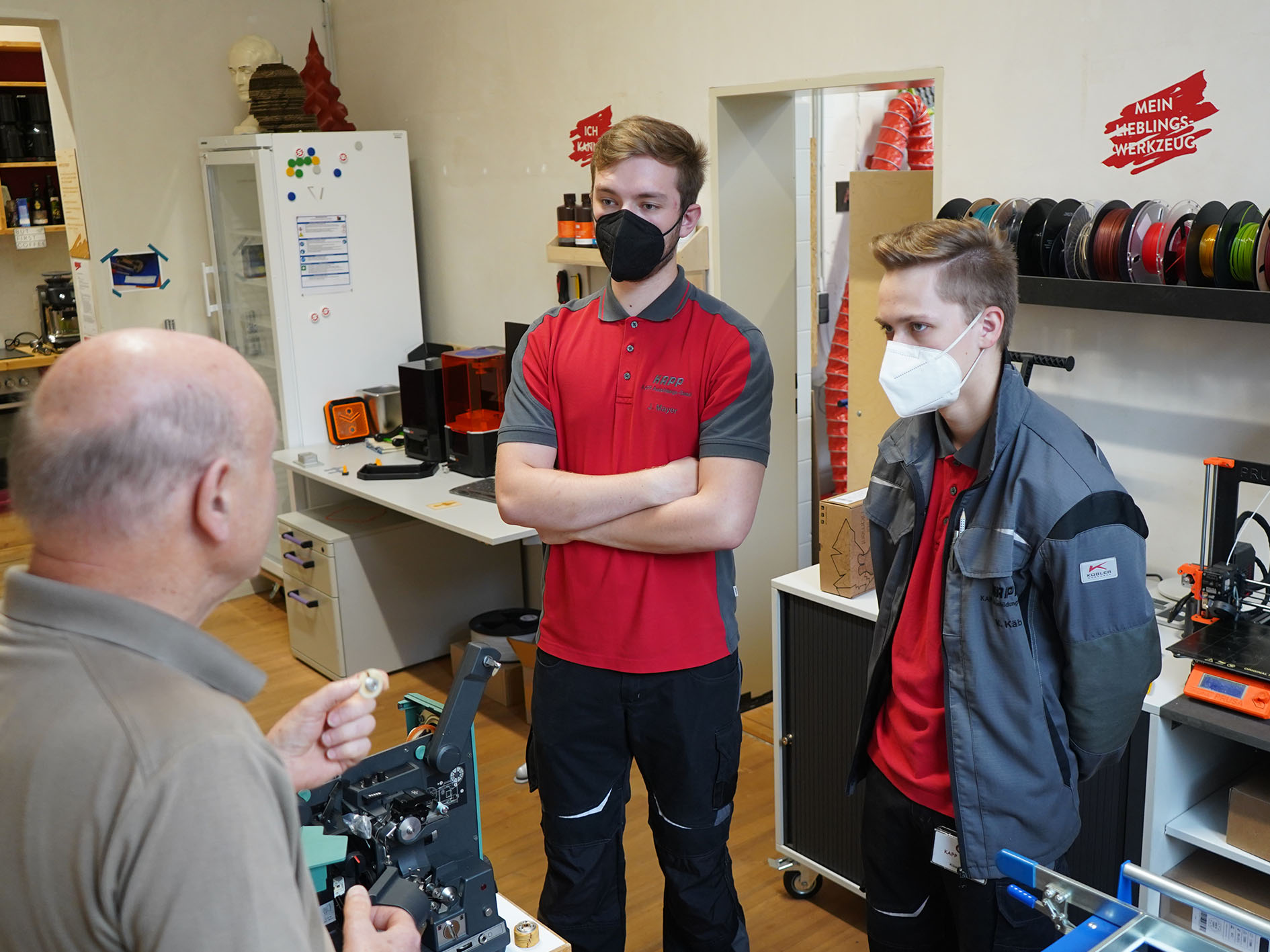 Drei Personen befinden sich in einer Werkstatt der Hochschule Coburg. Zwei junge Männer in passenden Uniformen und Masken stehen da und hören einem älteren Mann zu, der etwas mit einer Maschine vorführt. Der Raum ist voll mit Regalen voller Ausrüstung und Filamentspulen für 3D-Drucker.