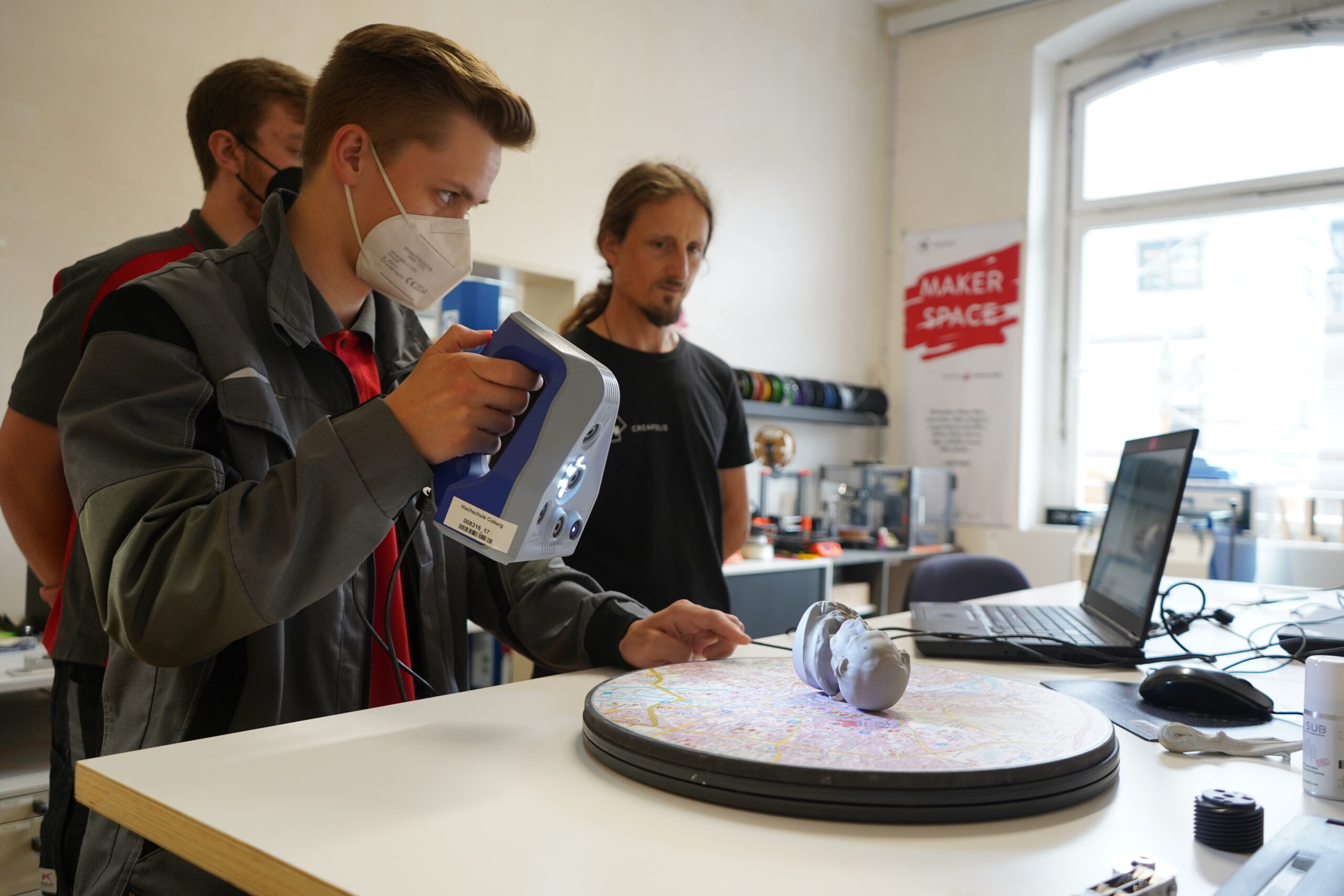 In einem lebendigen Makerspace der Hochschule Coburg bedient eine Person mit Maske gekonnt einen 3D-Scanner auf einer rotierenden Plattform, während eine andere den Vorgang beobachtet. Umgeben von Laptops und unterschiedlicher Ausrüstung verkörpern sie Innovation und Zusammenarbeit.