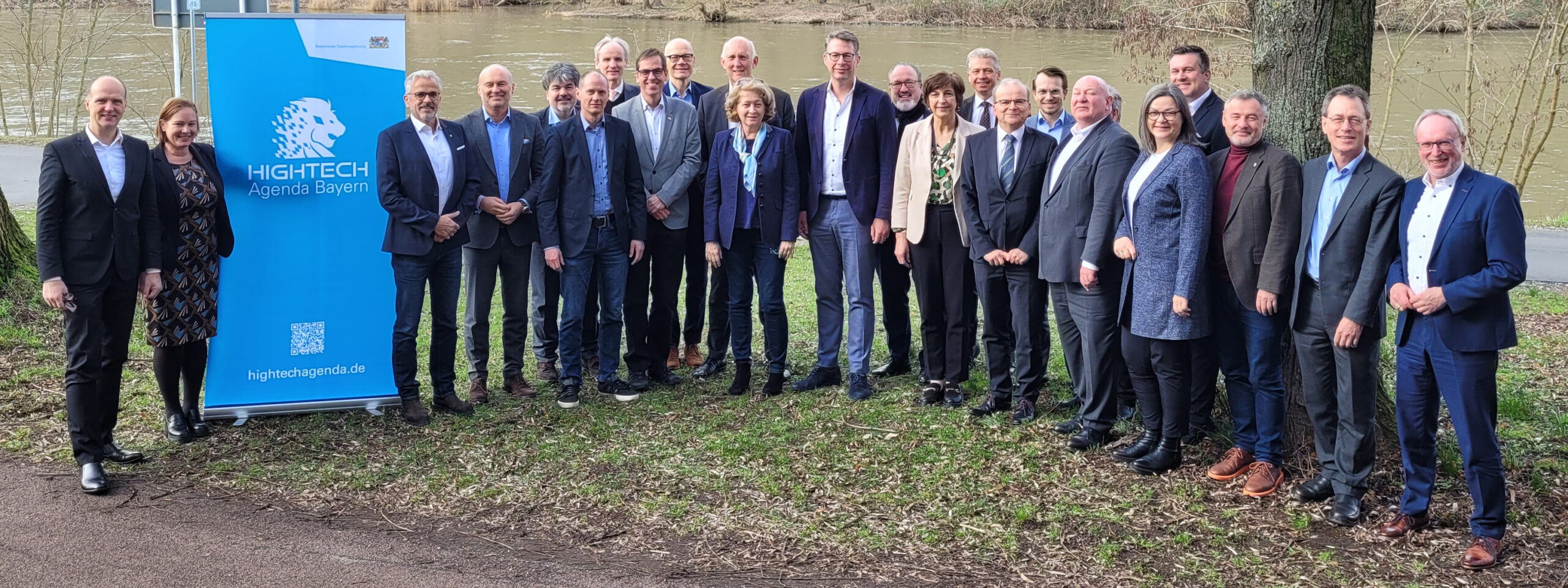 Eine Gruppe von Menschen in formeller Kleidung steht im Freien auf einer Wiese neben einem Fluss und posiert neben einem blauen Schild mit der Aufschrift „Hightech Agenda Bayern“. Mit Bäumen und dem Flussufer im Hintergrund verleihen Vertreter der Hochschule Coburg der Szene ihre Präsenz.