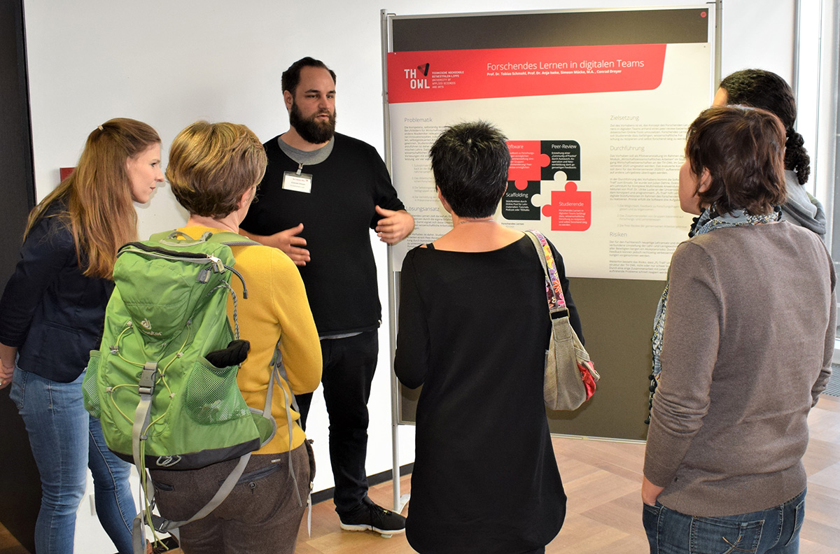 Eine Gruppe von Menschen steht im Halbkreis um einen Mann, der an der Hochschule Coburg ein Poster mit dem Titel „Forschendes Lernen in digitalen Teams“ präsentiert. Sie diskutieren in einem Innenraum. Der Mann gestikuliert, während er erklärt.