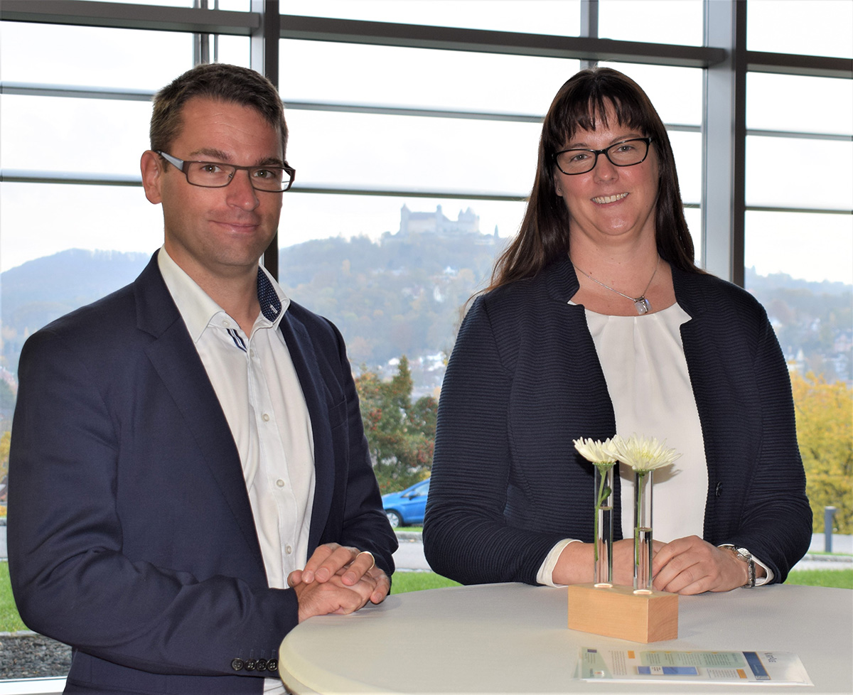 Ein Mann und eine Frau in Business-Kleidung stehen an einem runden Tisch mit zwei weißen Blumen in einer Vase. Sie lächeln herzlich und verkörpern die Professionalität, die mit der Hochschule Coburg verbunden wird. Hinter ihnen breitet sich durch ein großes Fenster eine malerische Hügellandschaft mit einem Schloss in der Ferne aus.