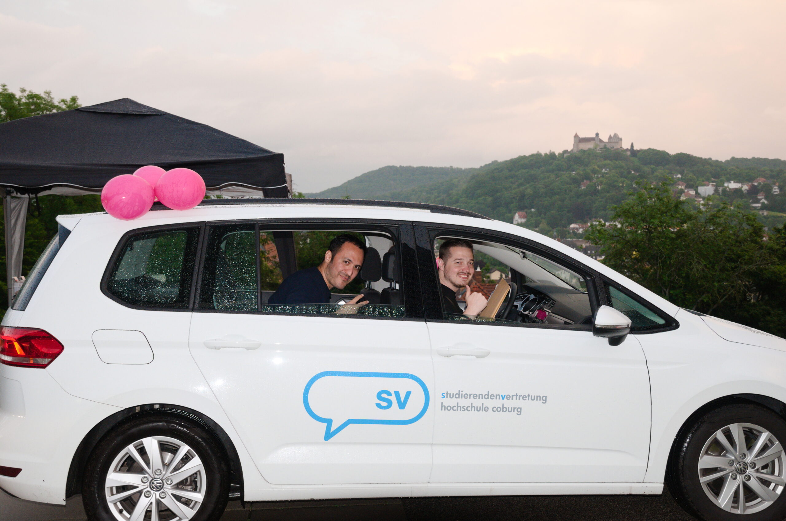 Zwei lächelnde Menschen sitzen in einem weißen Auto, das mit dem Logo der „Studierendenvertretung Hochschule Coburg“ geschmückt ist. Fröhlich schweben rosa Luftballons auf dem Dach. Dahinter sind ein malerischer Hügel und ein historisches Gebäude der Hochschule Coburg zu sehen, eingerahmt von einem wolkigen Himmel.