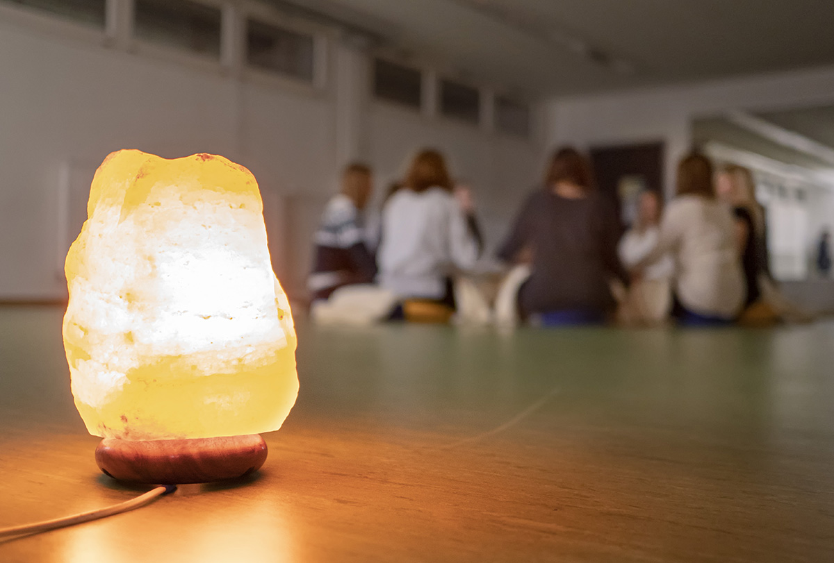 Im Vordergrund steht eine leuchtende Salzlampe auf einem Tisch. Im unscharfen Hintergrund sitzen Studierende der Hochschule Coburg im Kreis, was den Eindruck einer spannenden Gruppenaktivität oder Diskussion in einem schwach beleuchteten Raum erweckt.