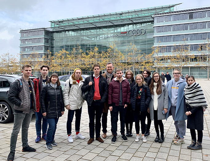 Eine Gruppe von Menschen, möglicherweise Studenten der Hochschule Coburg, steht vor dem Audi-Gebäude und lächelt in die Kamera. Die Kulisse ist eine moderne Plaza mit Bäumen und einer mehrstöckigen Glas- und Stahlkonstruktion im Hintergrund. Alle sind in Winterkleidung gekleidet.