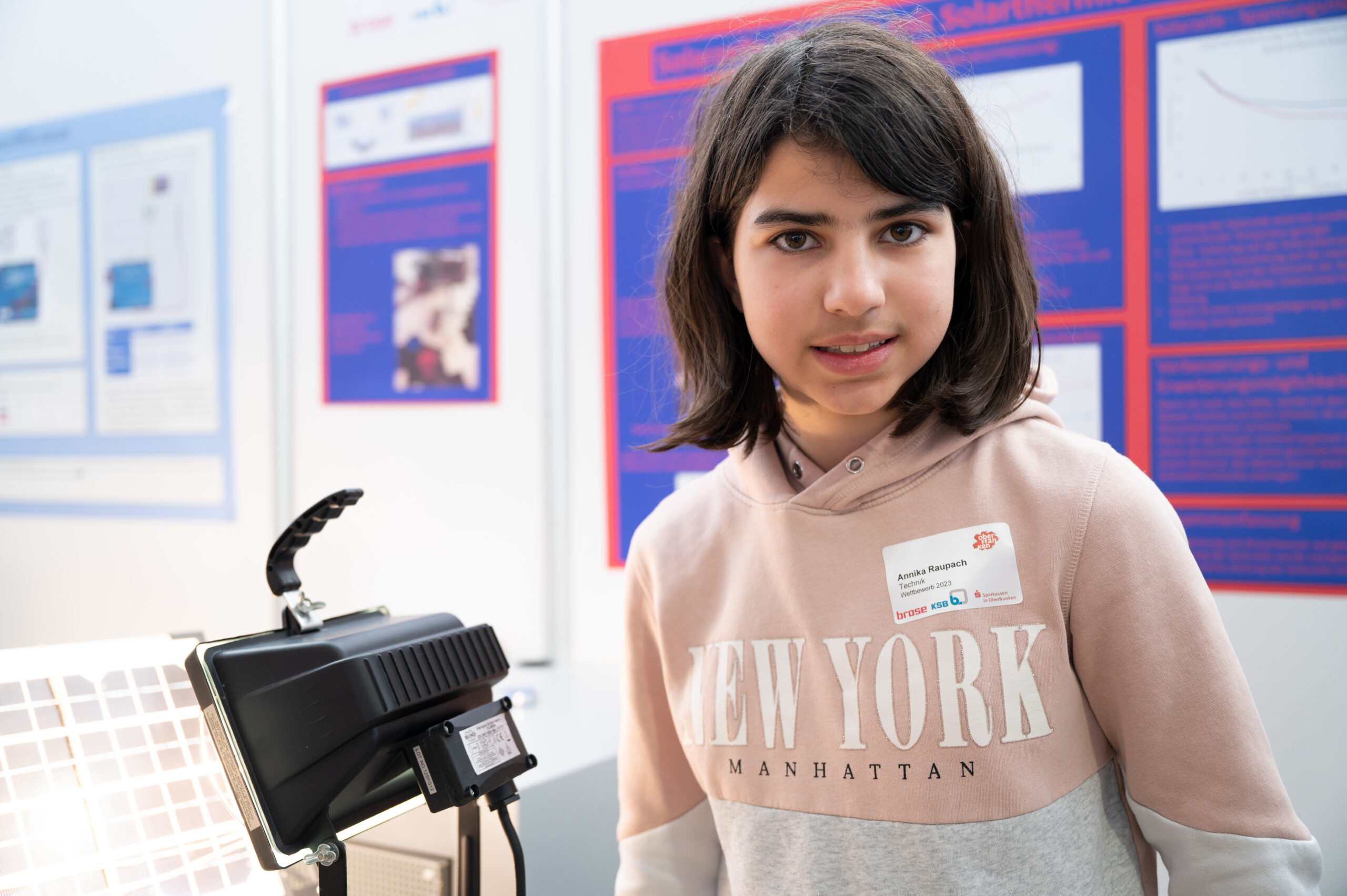 Ein junger Mensch mit dunklem Haar steht vor einer Reihe von Informationsplakaten. Er trägt ein hellrosa Sweatshirt mit der Aufschrift „New York“. Daneben befindet sich eine wissenschaftliche Präsentation mit Lichtern und einem kleinen Gitter, die vermutlich Innovationen der Hochschule Coburg präsentiert.