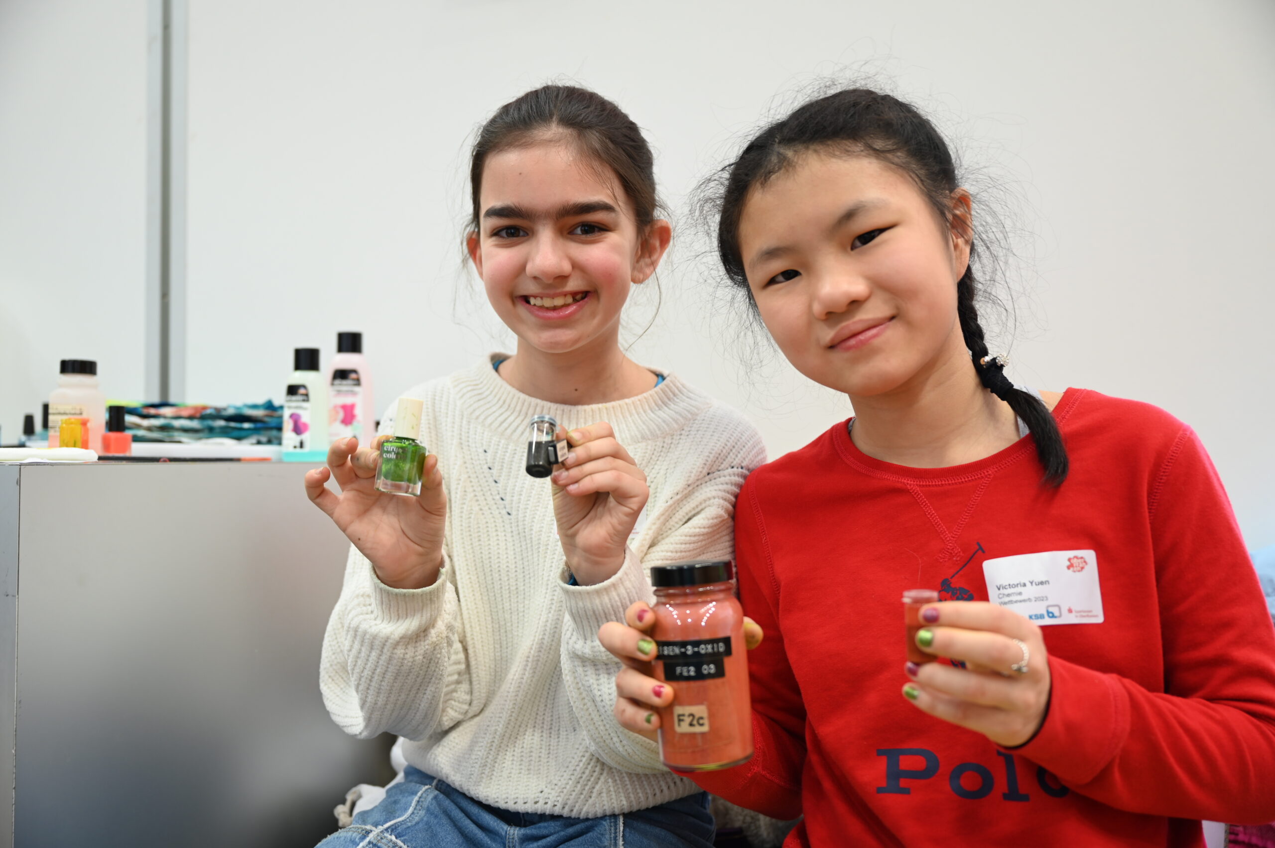 Zwei junge Mädchen lächeln in die Kamera. Jedes hält kleine Gläser mit buntem Inhalt in den Händen. Lässig gekleidet sitzen sie in einer Werkstatt, die an die Hochschule Coburg erinnert, und strahlen Freude und Neugier aus. Im Hintergrund sind noch ein paar Gläser und Flaschen zu sehen.