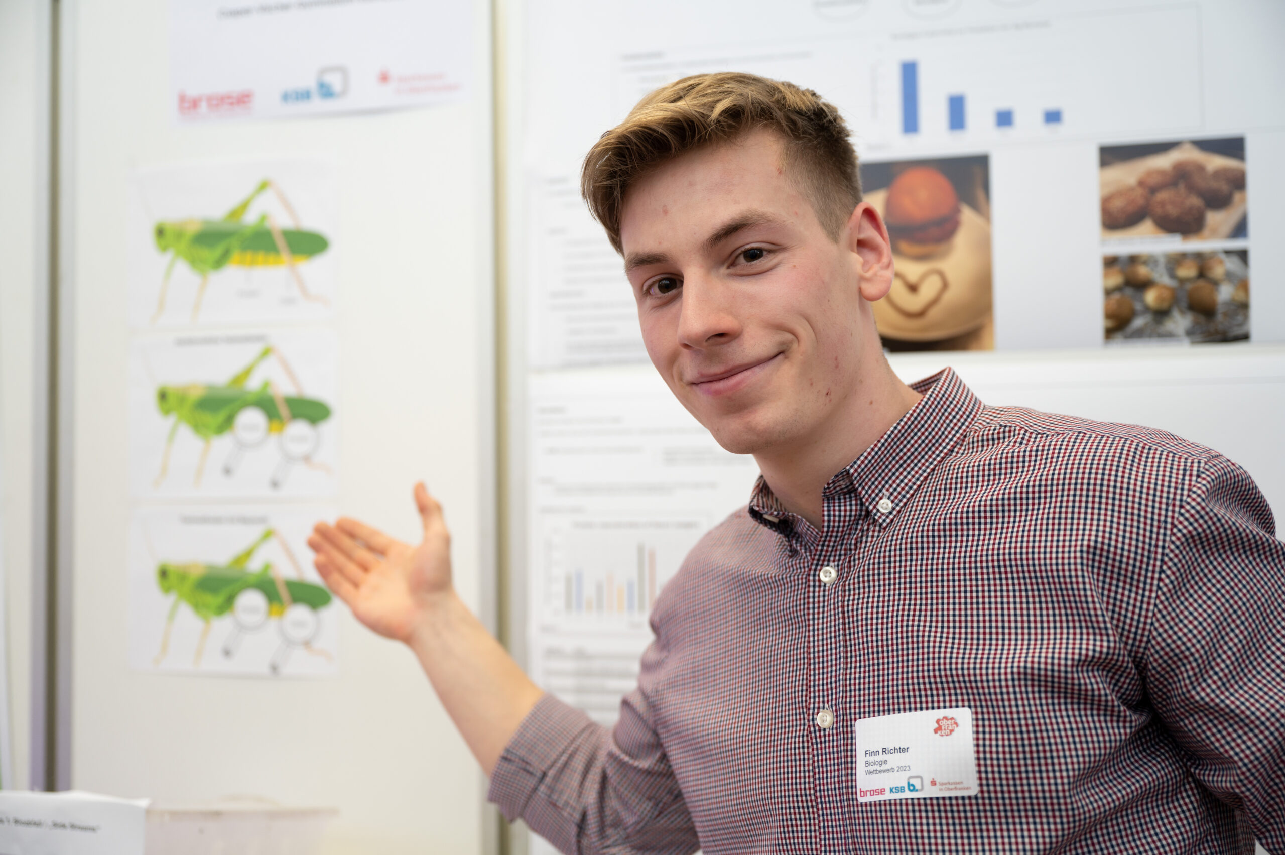 Eine Person steht vor einer wissenschaftlichen Präsentationstafel, lächelt und zeigt Bilder einer Heuschrecke und verschiedene Diagramme. Auf der Tafel sind Diagramme und Lebensmittelfotos zu sehen. Sie trägt ein kariertes Hemd und ein Namensschild der Hochschule Coburg und präsentiert stolz ihre Erkenntnisse.