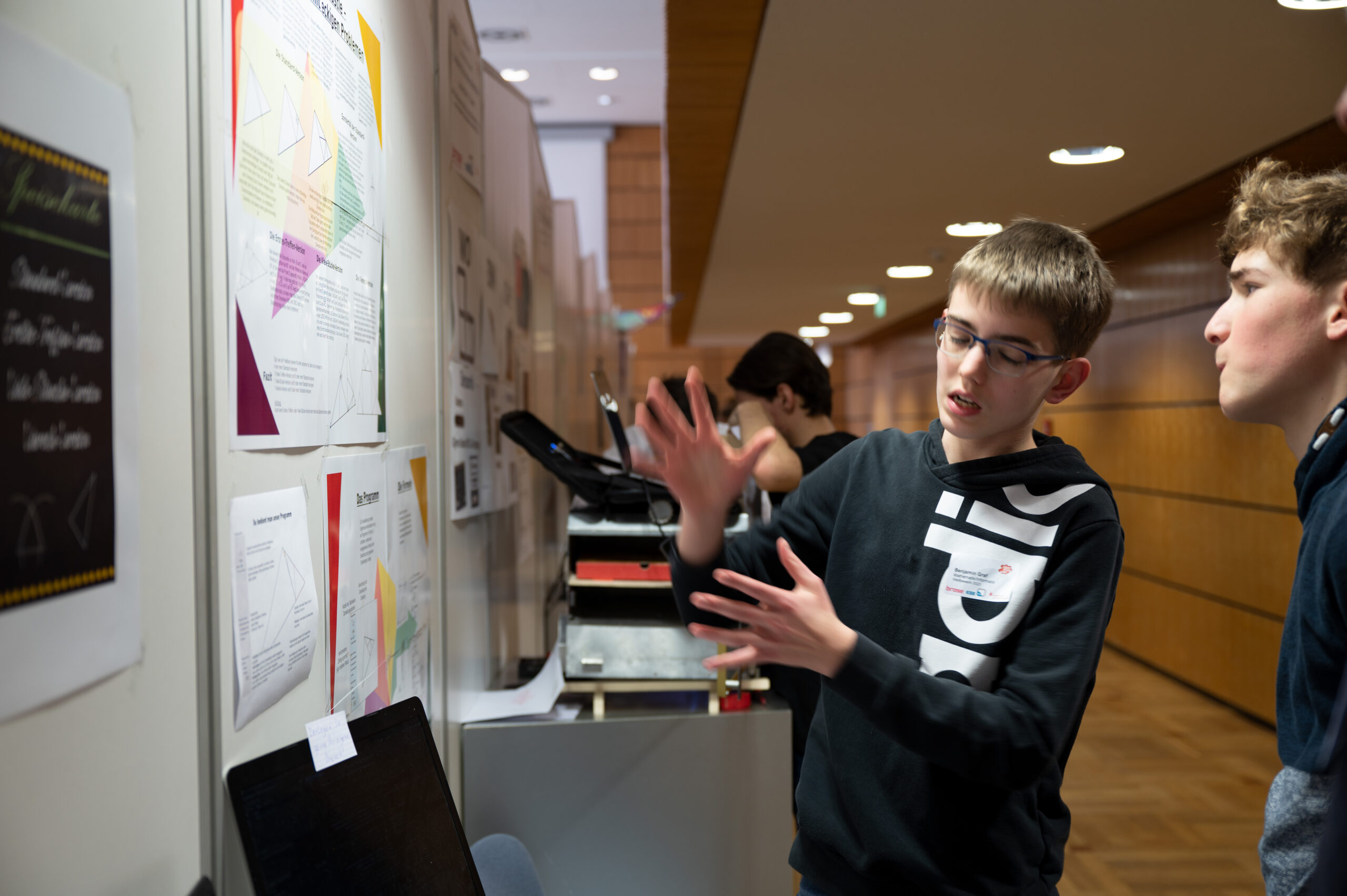 In einem belebten Flur der Hochschule Coburg erklärt ein junger Mensch einer anderen Person lebhaft farbenfrohe Poster mit bunten Diagrammen und Texten. In der Nähe steht jemand mit einem offenen Ordner, der leicht verschwommen im Hintergrund zu sehen ist, und ist völlig in seine Studien vertieft.