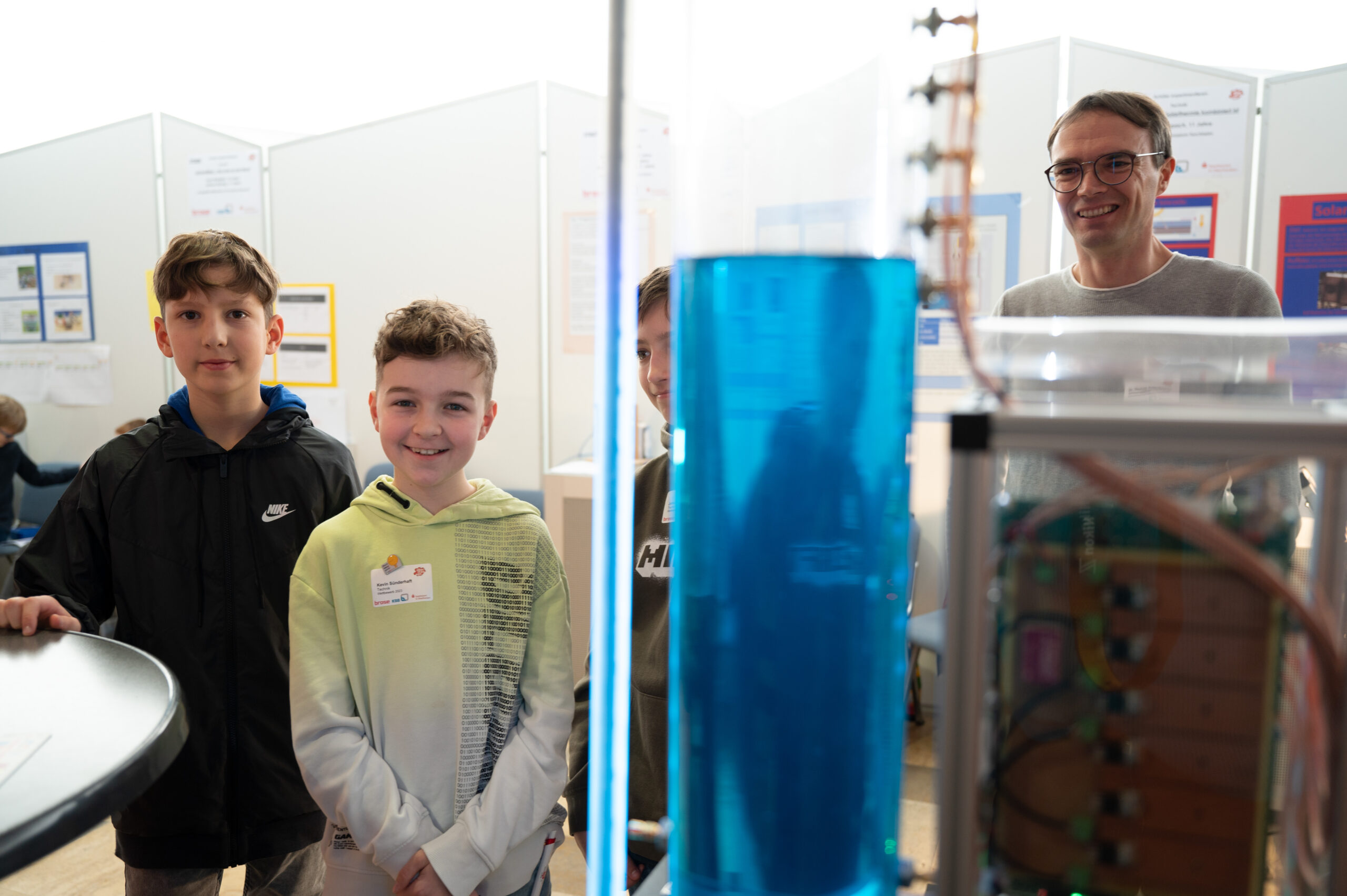 Eine Gruppe von drei Kindern und einem Erwachsenen steht lächelnd in einem naturwissenschaftlichen Ausstellungsraum der Hochschule Coburg. Im Vordergrund ist ein hoher, durchsichtiger Zylinder zu sehen, der mit einer blauen Flüssigkeit gefüllt ist.