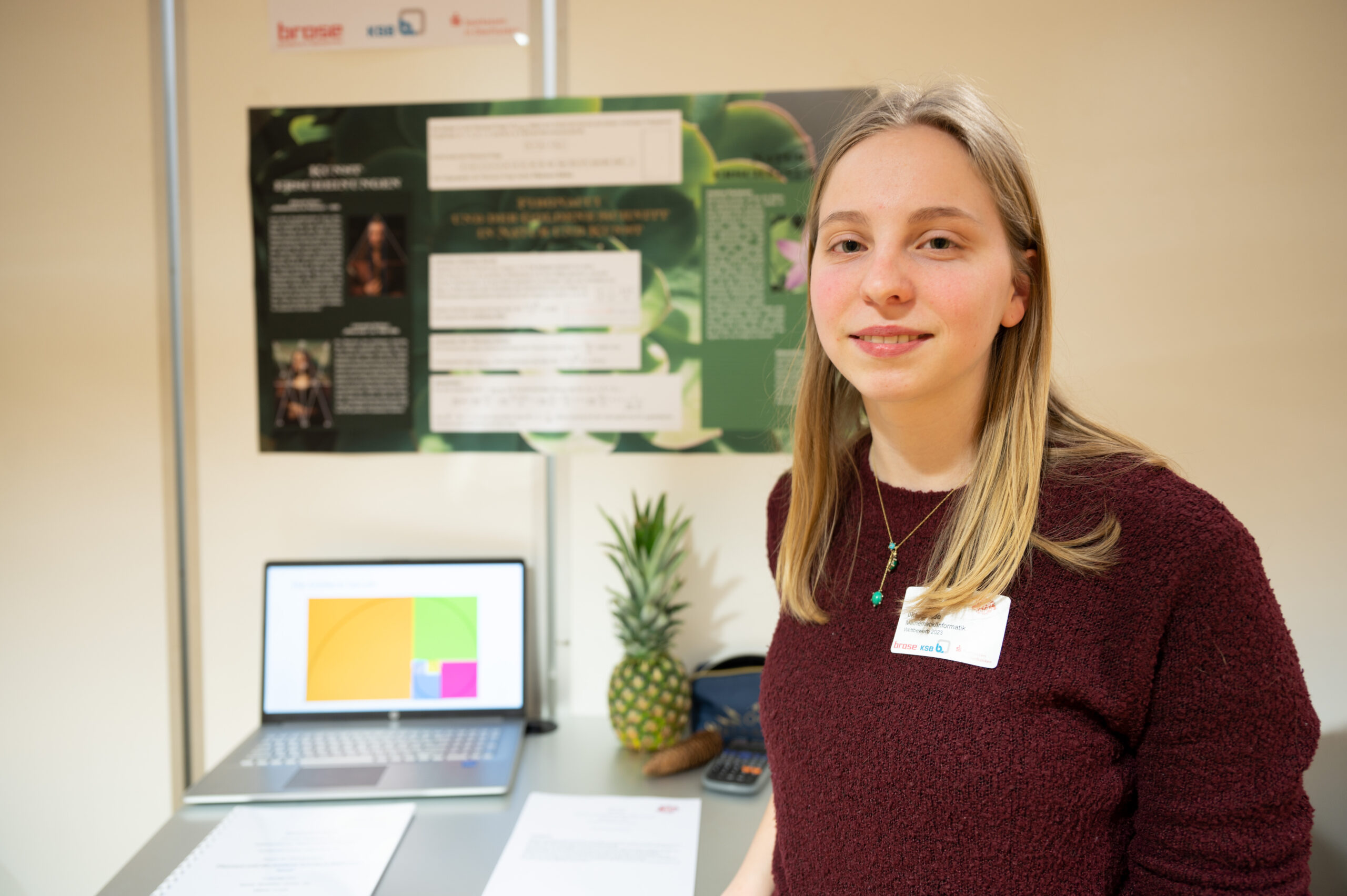 Eine Person steht vor einem Ausstellungsstand mit einem Laptop, einer Ananas und einem Informationsplakat der Hochschule Coburg. Sie lächelt und trägt einen kastanienbraunen Pullover und ein Namensschild. Der Stand scheint Teil einer Präsentation oder Ausstellung zu sein.