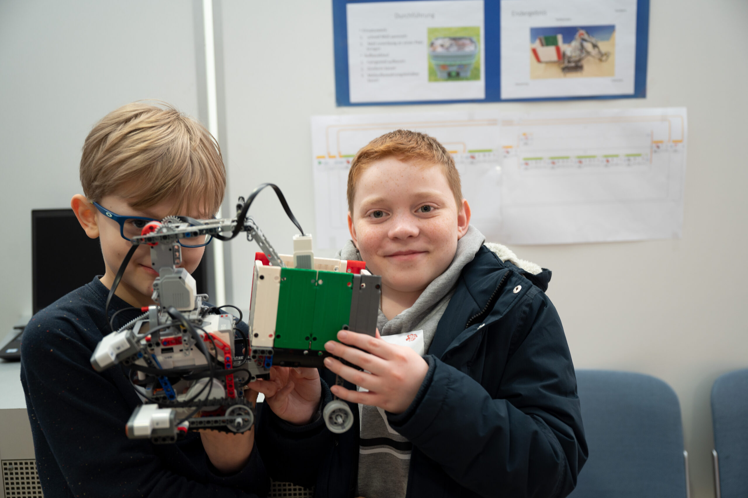 In einem Klassenzimmer der Hochschule Coburg lächeln zwei Kinder und halten ein Robotergerät in der Hand. Eines trägt eine Brille, das andere hat kurze rote Haare. Hinter ihnen deuten Diagramme und ein Computer auf dem Tisch auf ihre innovative Lernumgebung hin.