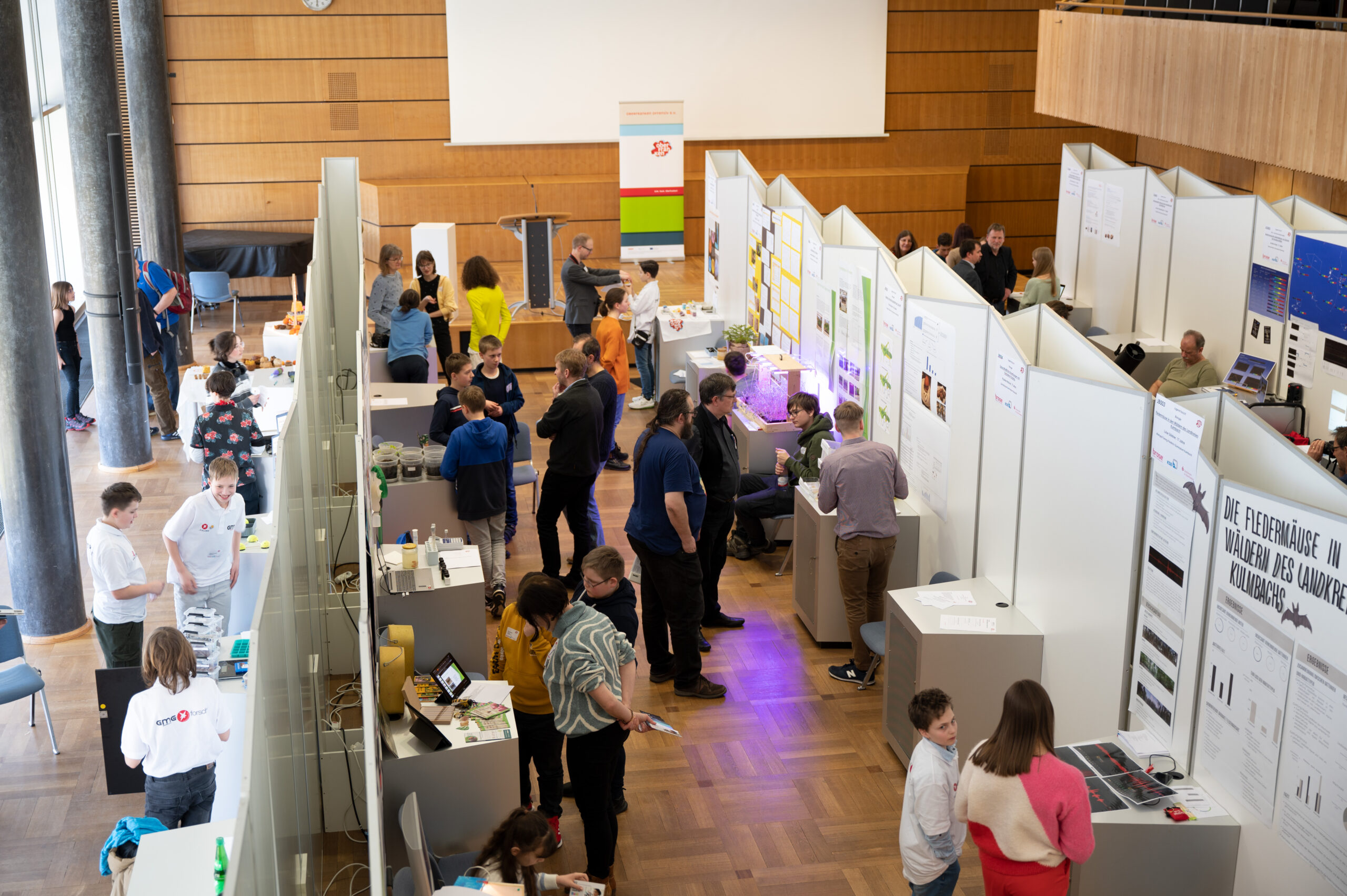 Die große Indoor-Wissenschaftsmesse der Hochschule Coburg bietet eine Vielzahl von Ständen. Menschen jeden Alters beteiligen sich an lebhaften Diskussionen und Vorführungen. Poster und Schautafeln säumen die Trennwände, während natürliches Licht den lebendigen Raum erfüllt.