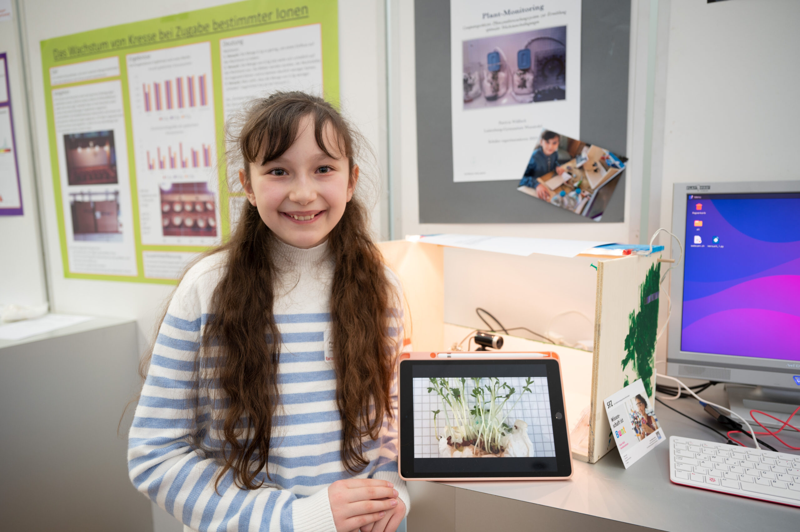 Ein junges Mädchen mit langen braunen Haaren und einem gestreiften Pullover steht lächelnd neben ihrem Wissenschaftsprojekt der Hochschule Coburg. Das Projekt beschäftigt sich mit der Überwachung von Pflanzen und wird auf einem Tablet angezeigt. Im Hintergrund sind Lehrplakate und ein digitaler Bildschirm zu sehen.