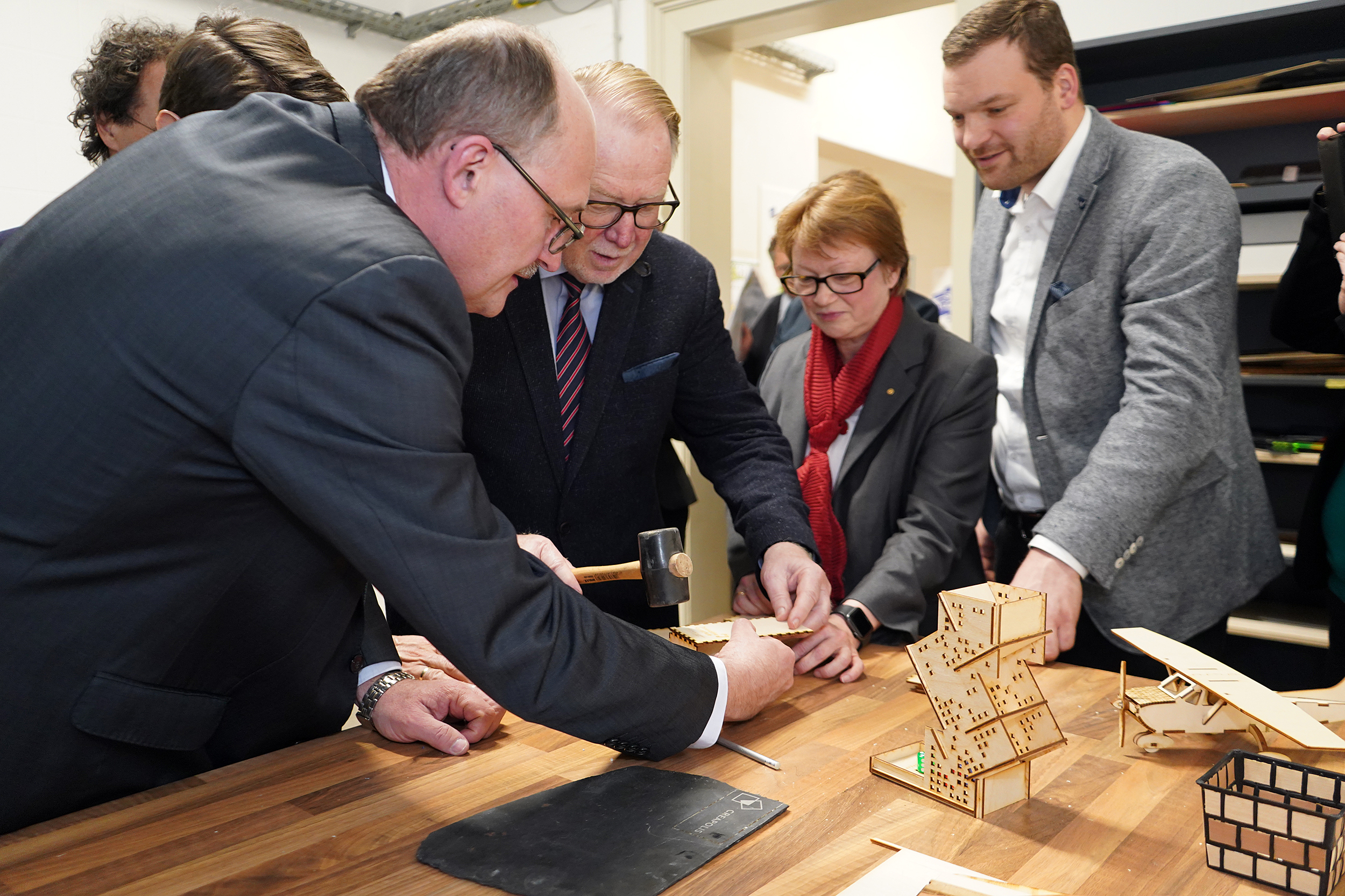 An der Hochschule Coburg versammelt sich eine Gruppe von Menschen in formeller Kleidung um einen Tisch und bastelt kleine Holzkonstruktionen. Ein Mann benutzt einen Hammer, um die Teile zusammenzusetzen, während andere zuschauen und assistieren. Der Tisch ist mit Modellteilen und Werkzeugen übersät, was Teamarbeit und Kreativität in einem akademischen Umfeld fördert.