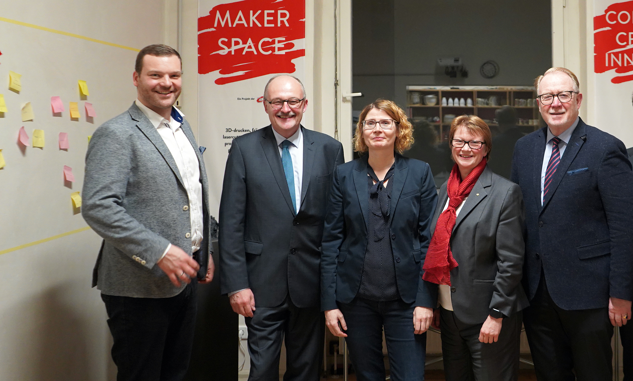 Fünf Personen stehen in einem Raum mit „Maker Space“-Schildern an den Wänden der Hochschule Coburg. Sie sind in Business-Kleidung gekleidet und lächeln in die Kamera, während bunte Haftnotizen den Hintergrund schmücken.